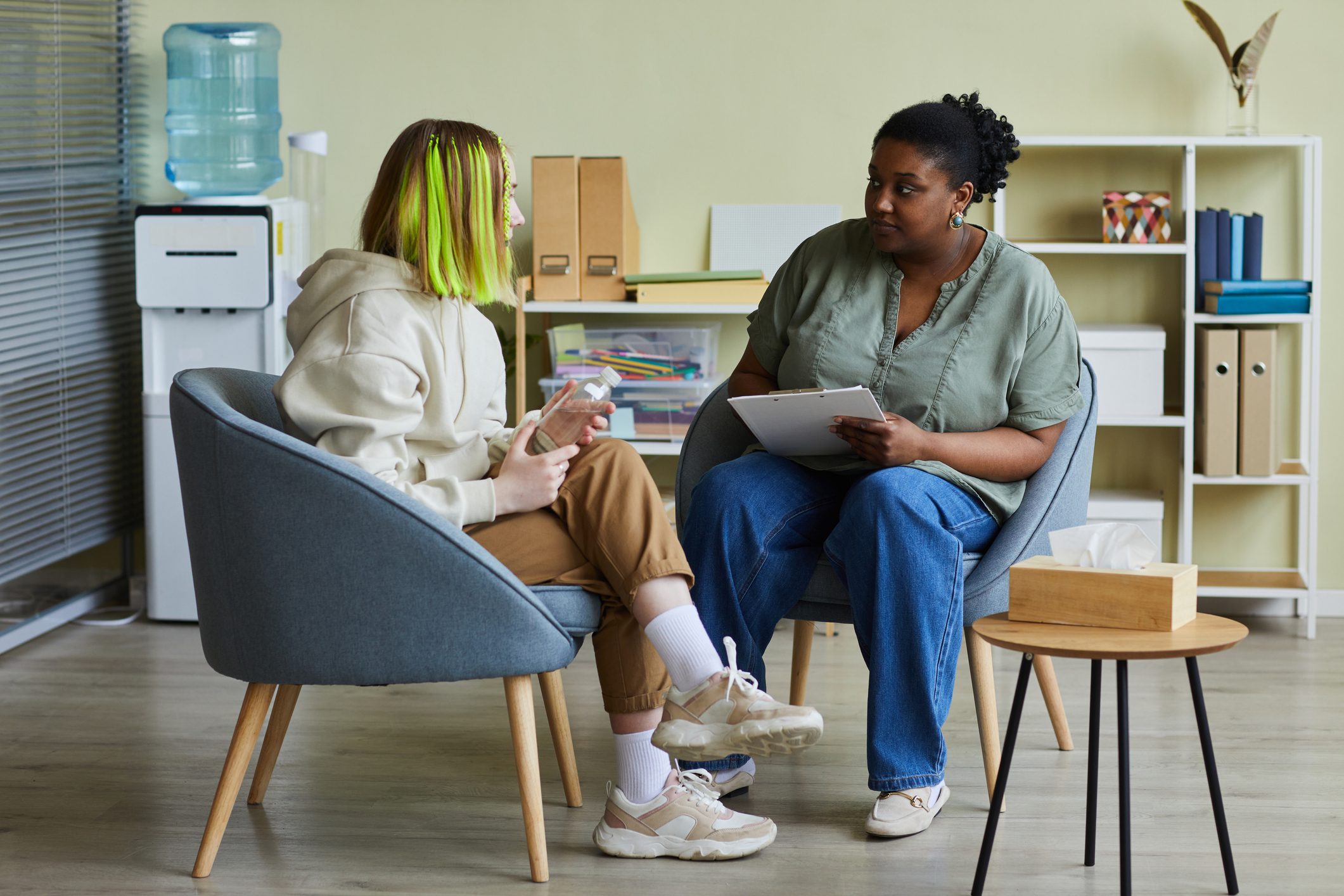 A Black, female psychologist sitting in a nicely decorated office starting treatment for an anxious teenage girl with bright green extensions in her hair
