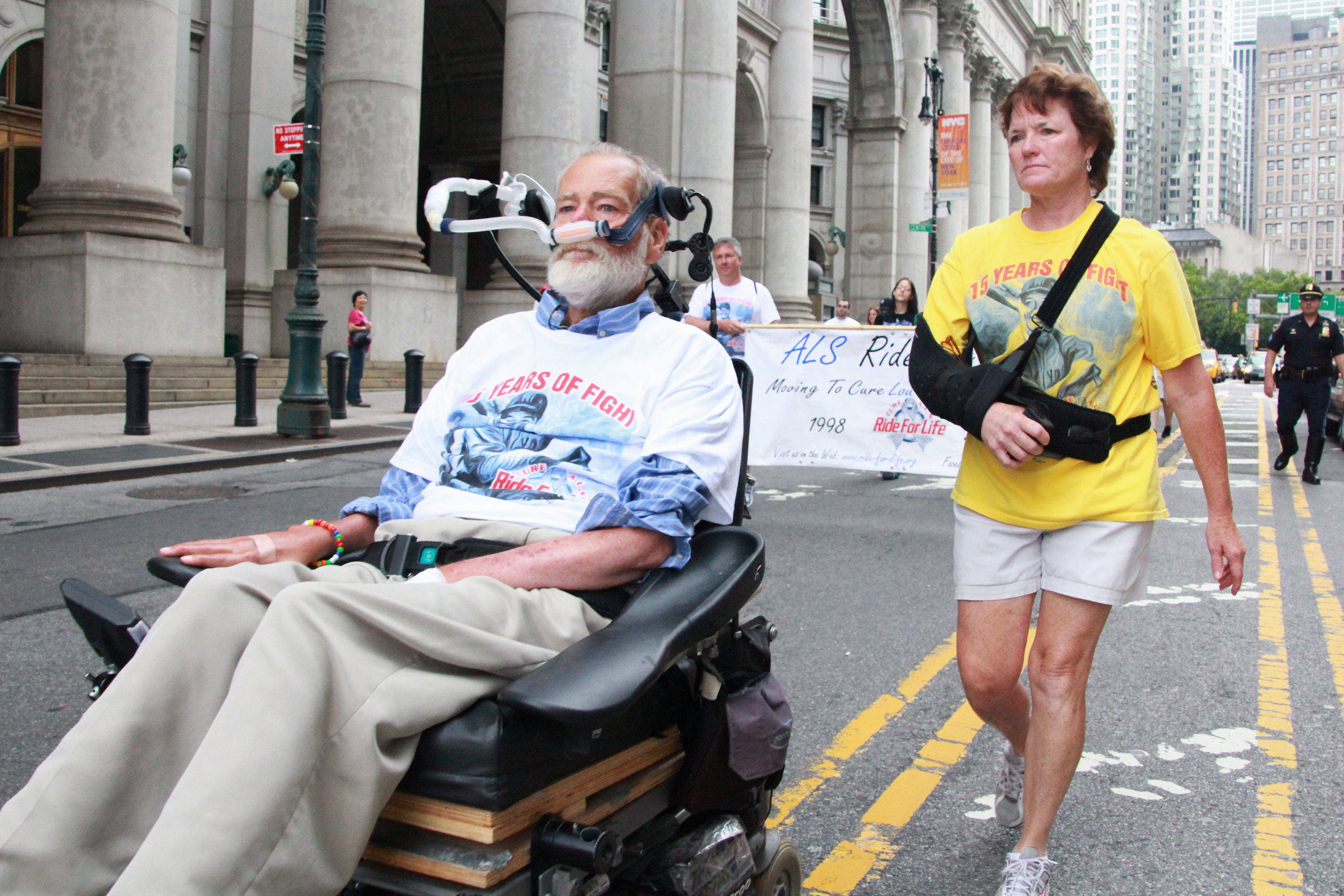  A man in a motorized wheelchair and a woman with her arm in a sling leading a charity walk for ALS research down a city street