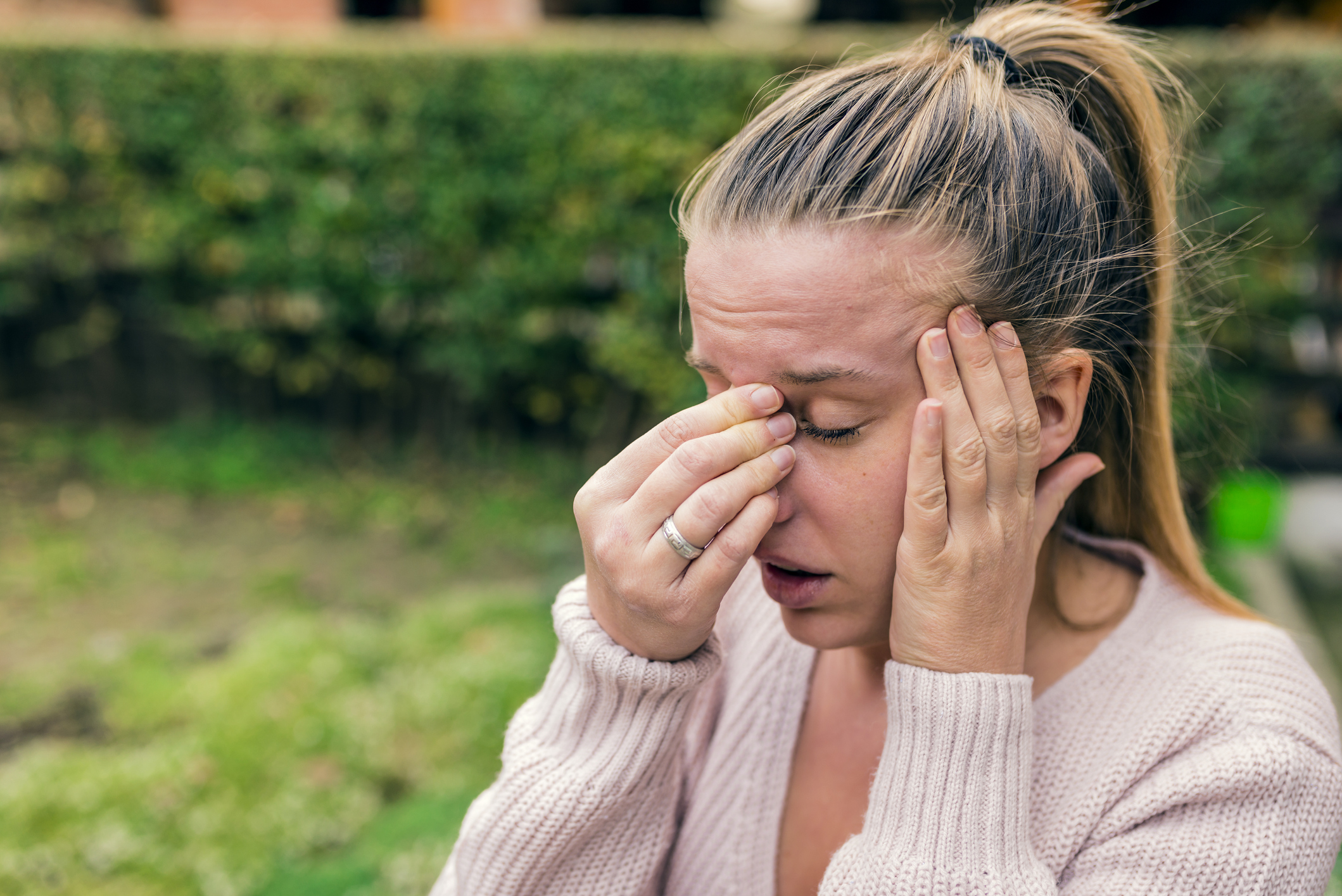 A flushed, dizzy white woman with a blonde ponytail holds her head in pain in her front yard