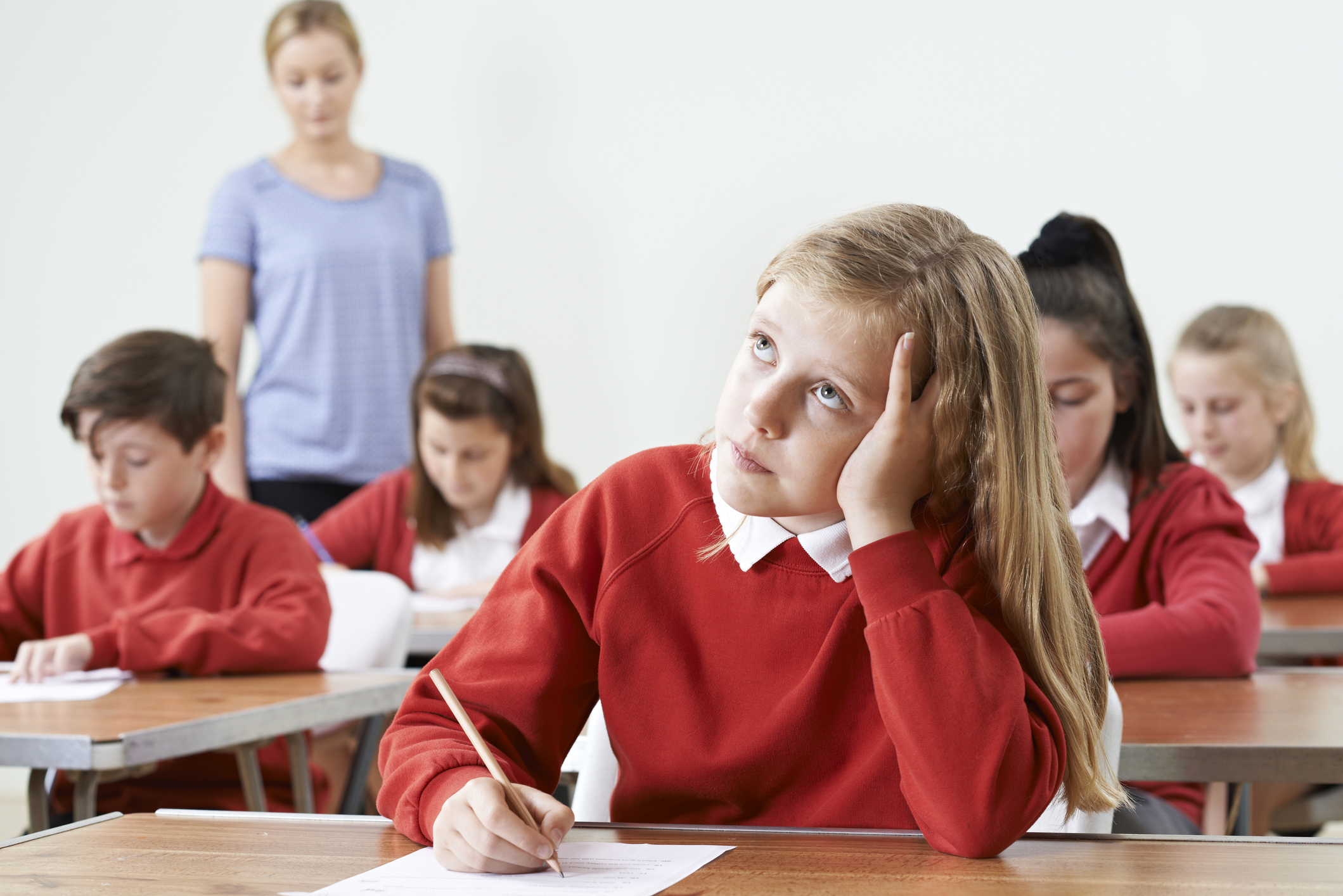 Middle school students in red school uniforms work on a class assignment while one blonde girl sits confused, not understanding the instructions