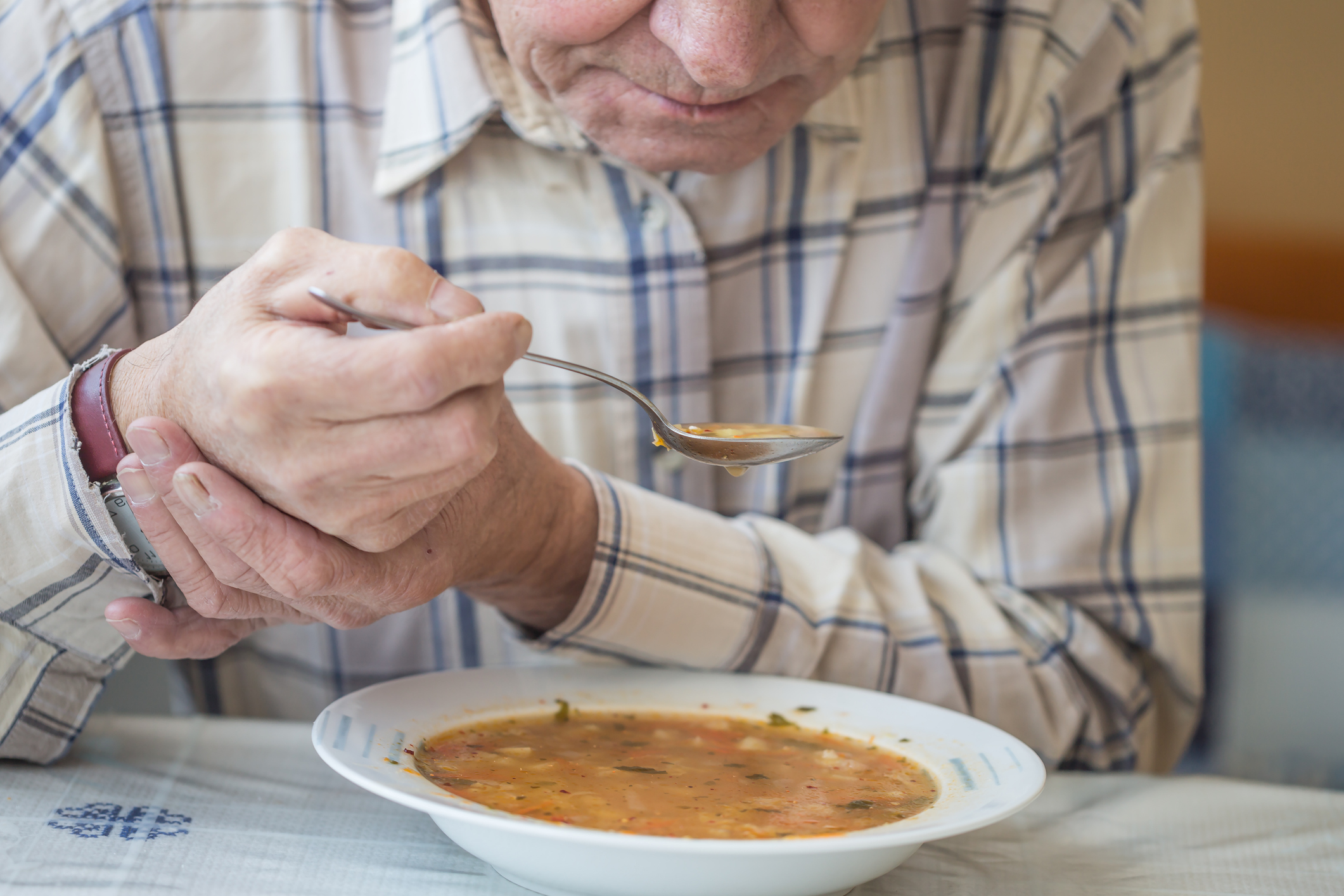  A elderly white man in a white plaid shirt seated at a dining table holds his wrist to stabilize his hand as he lifts a spoonful of soup to his mouth