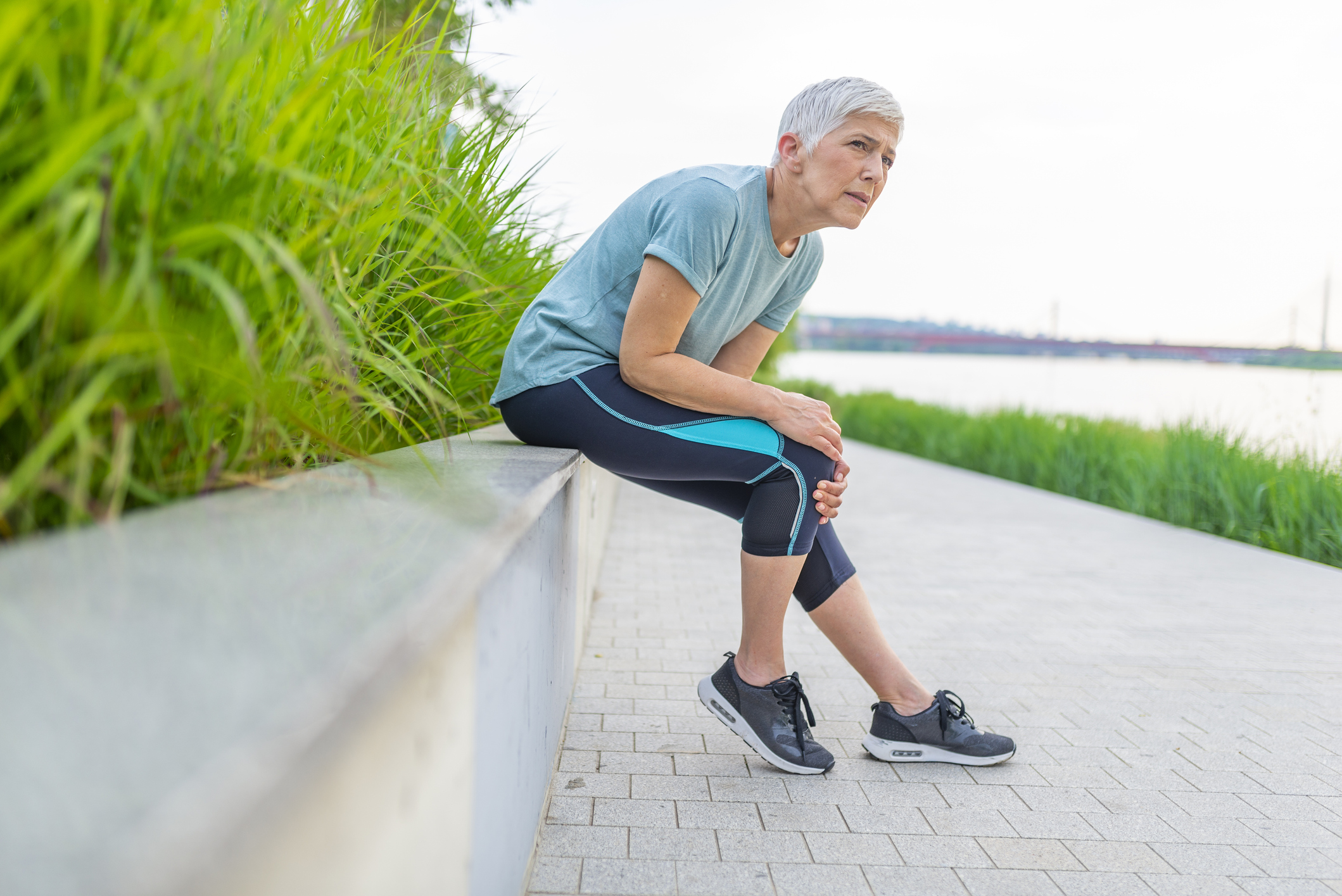 A middle aged white woman with short white hair and blue exercise clothes interrupts her run along a river to sit down and massage her knee