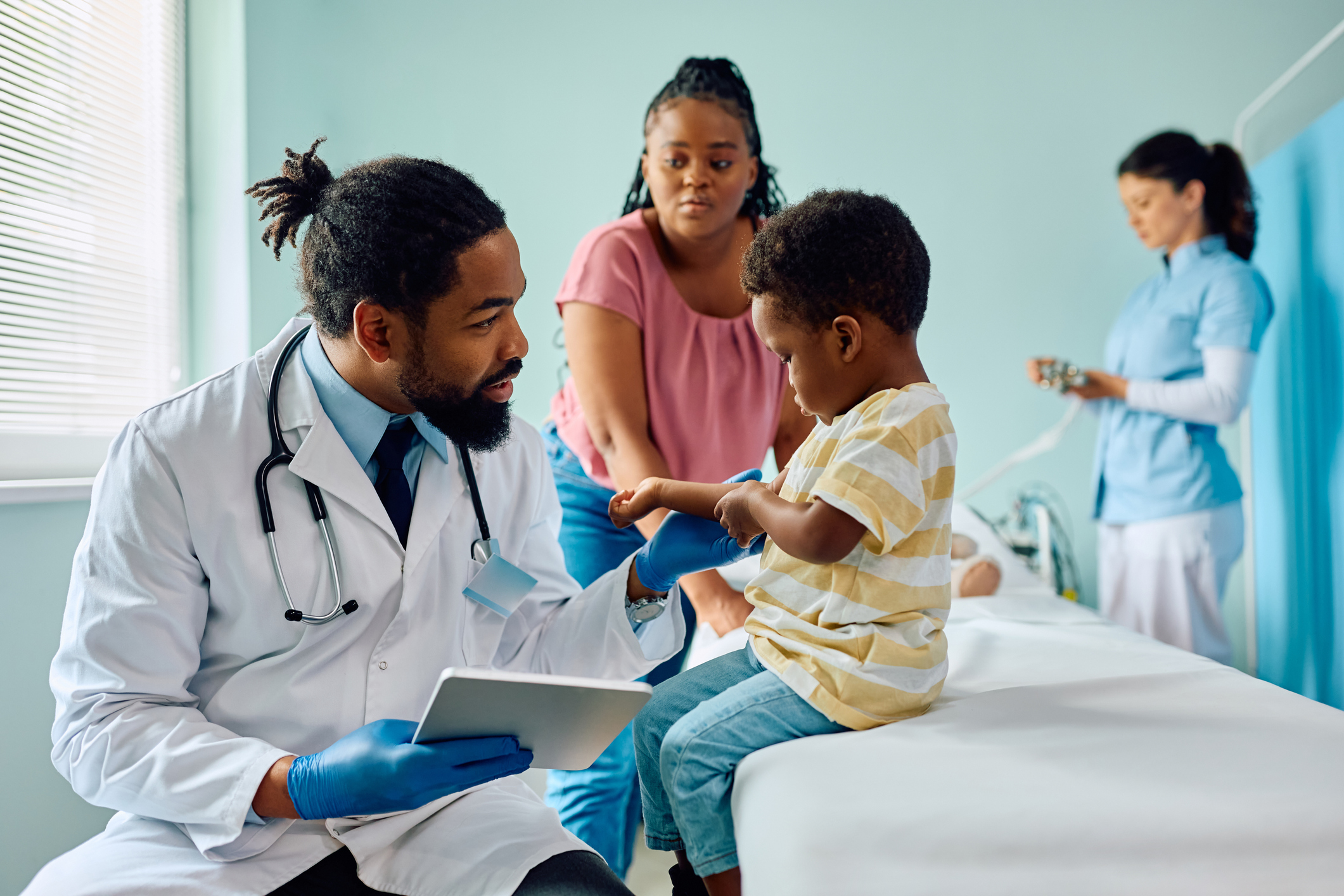 A young, male African American doctor with a beard and a short ponytail checks a young Black boy's joints for mobility issues during a regular sickle cell disease checkup while the boy's mother, a young Black woman with her hair in a braided ponytail, watches from a distance and a female nurse in blue scrubs takes notes.