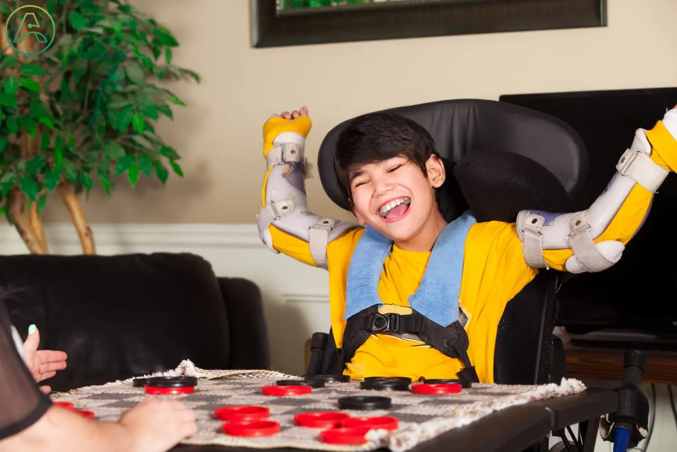 Young Asian boy in a wheelchair and arm braces sits at a table playing a game.