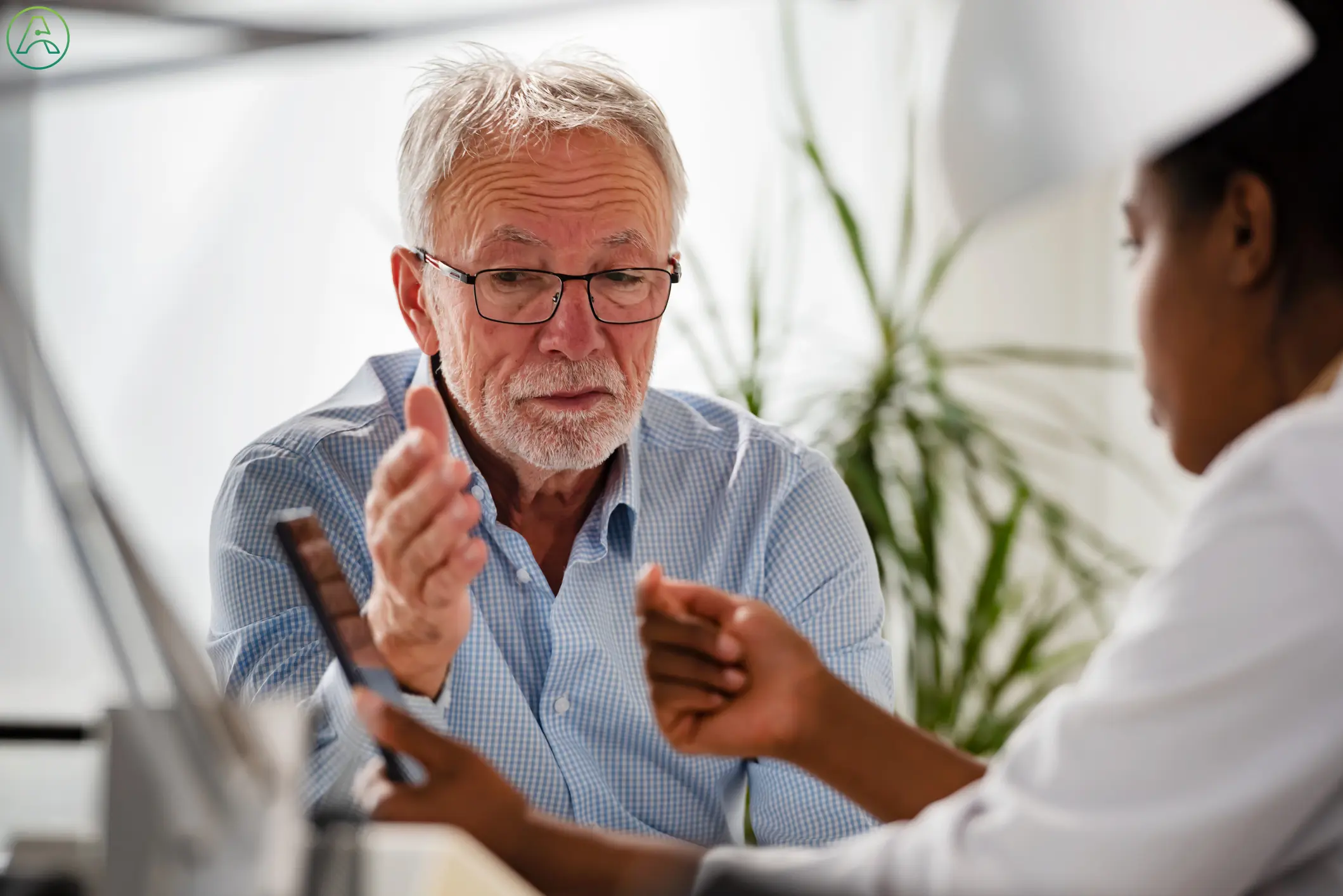 A Black female doctor explains smartphone accessibility functions to a senior man with white hair and a blue checkered shirt, who is frustrated by communication disabilities.