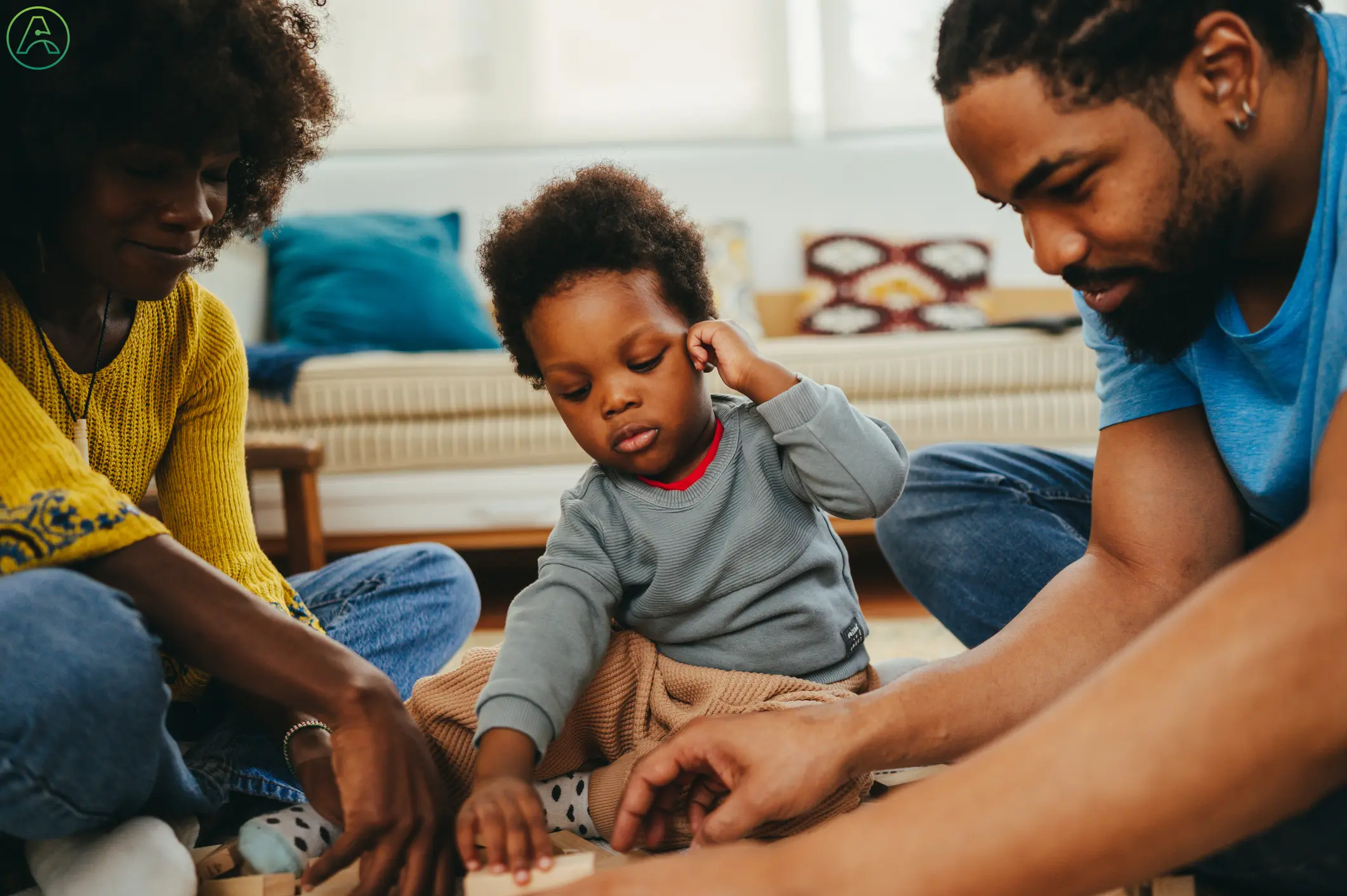 A Black toddler boy in a gray sweater and corduroy pants tugs on his ear while his parents teach him to stack blocks.