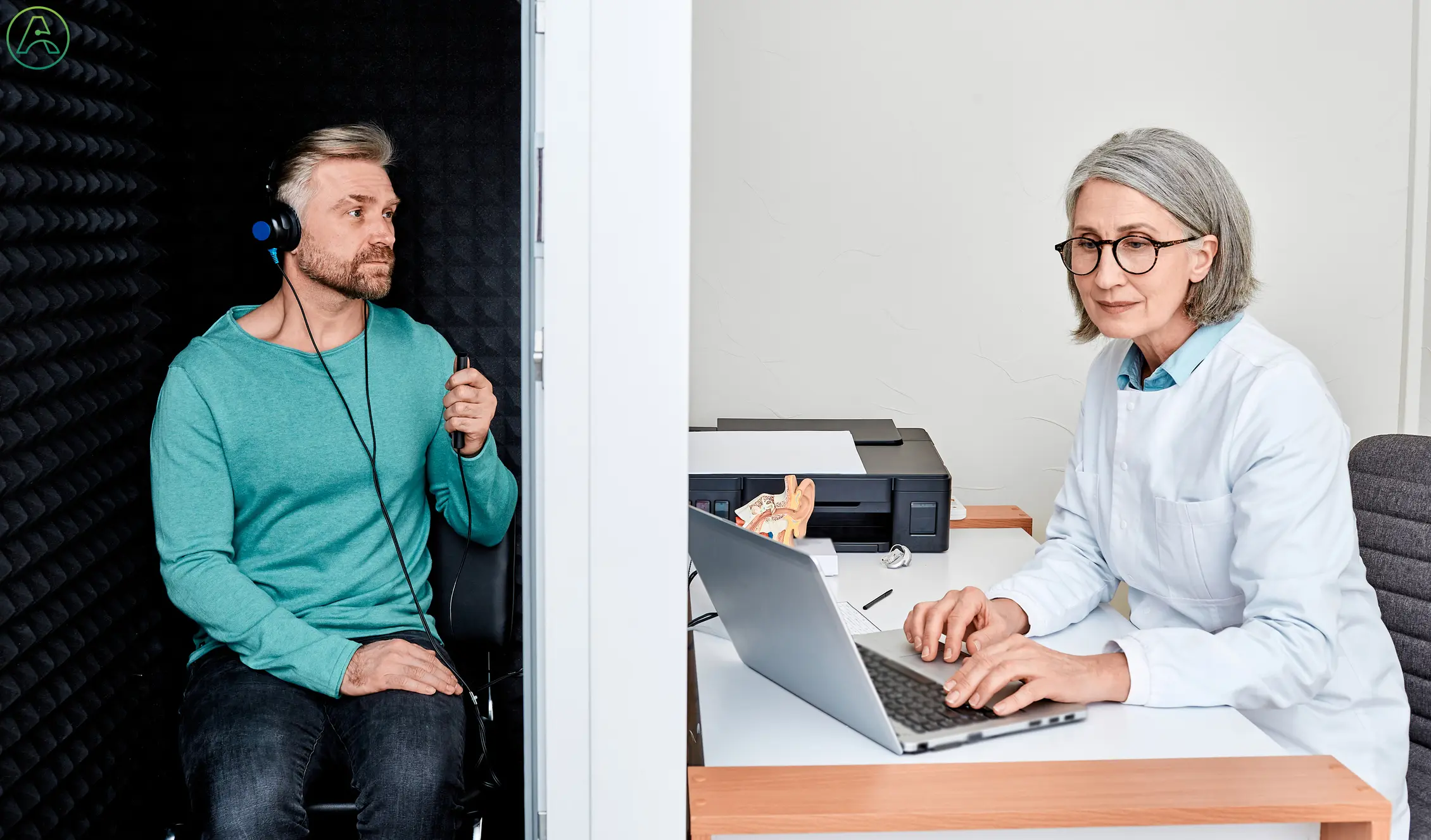 A white female audiologist sits at a desk reading results on a laptop while a white man in a blue shirt and jeans sits in the sound booth, taking a hearing test for tinnitus.