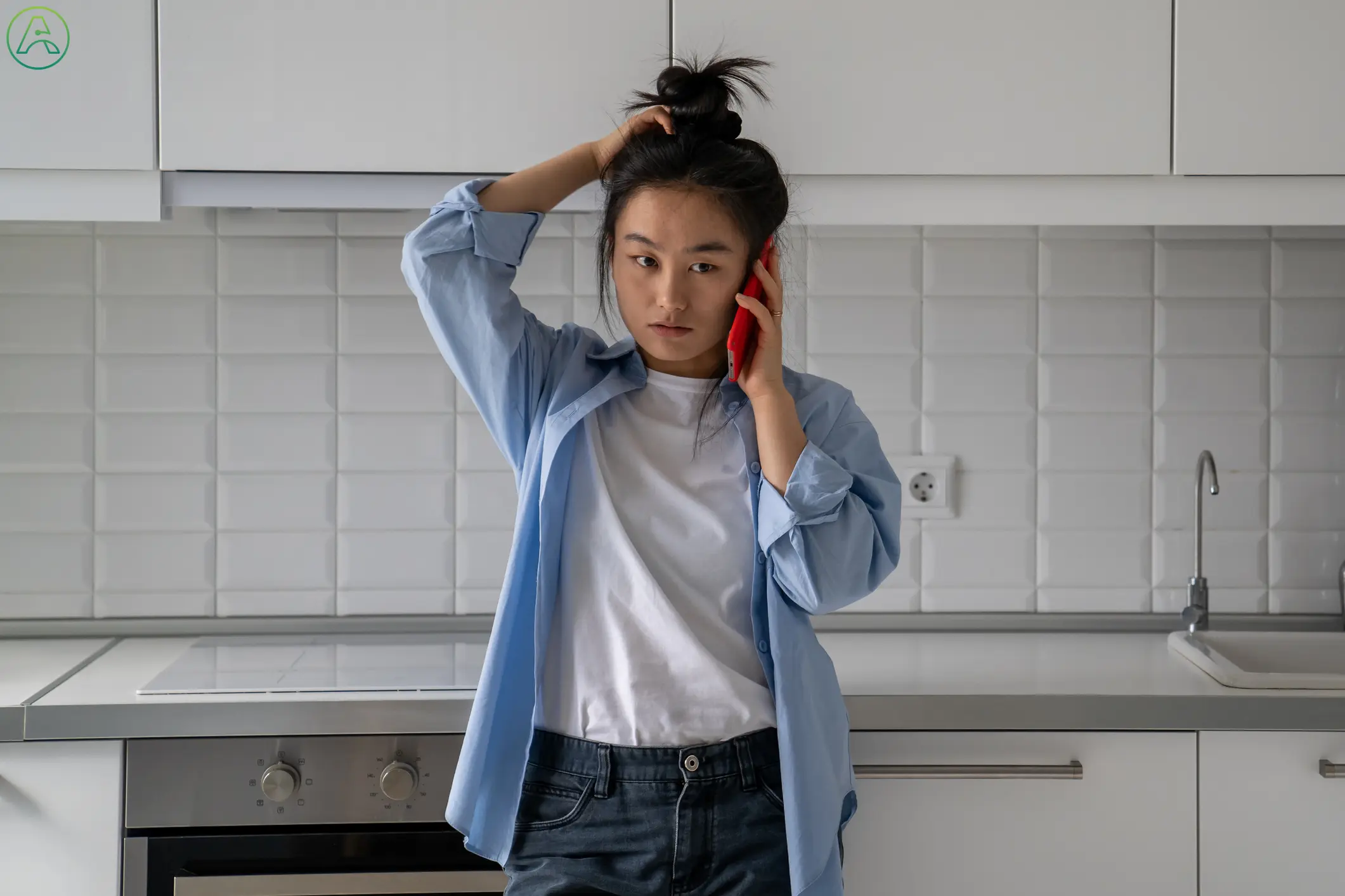 A frustrated young woman rubs her head while standing anxiously in her kitchen, trying to understand what someone says over the phone.