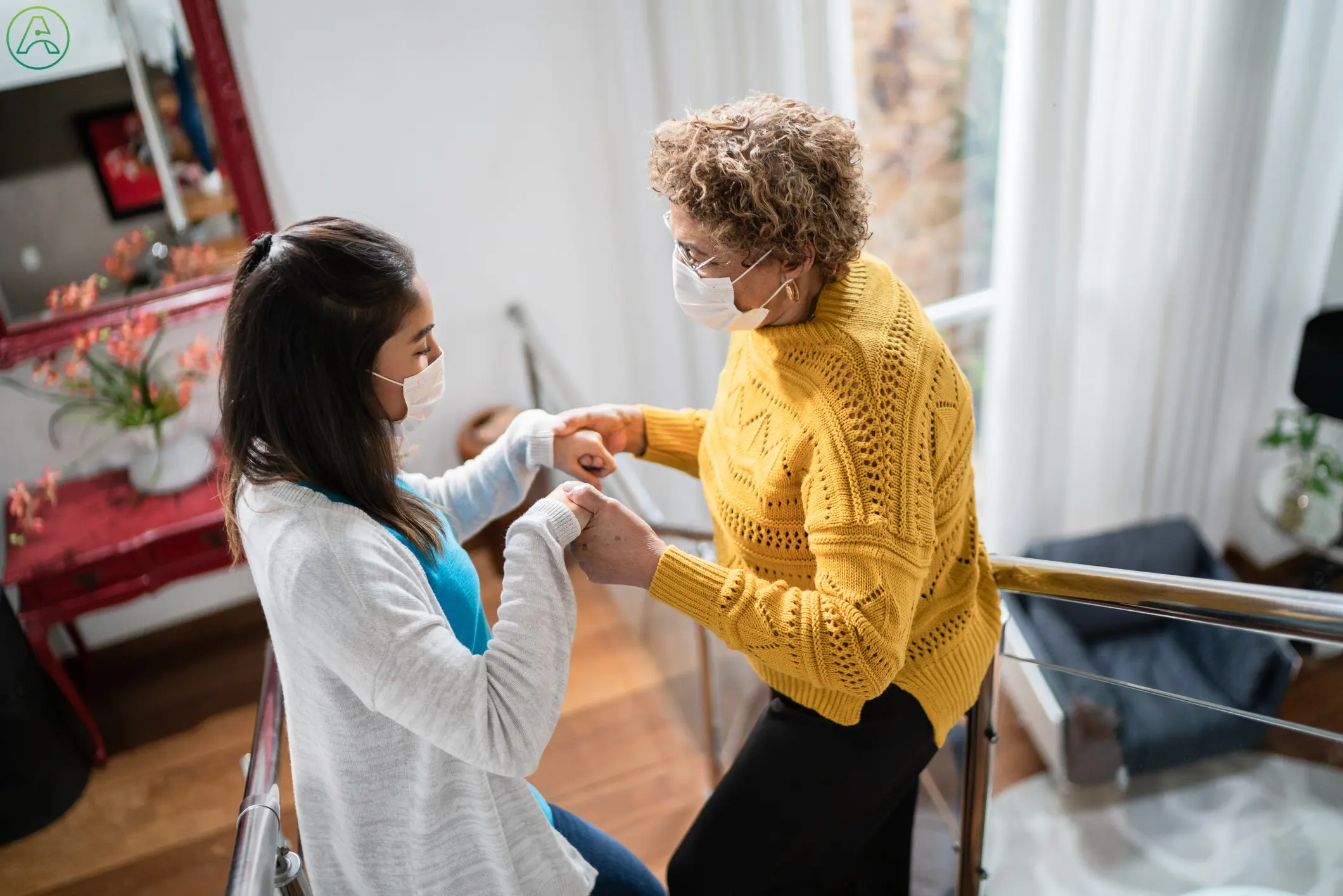 A Latina woman in a white cardigan and a white medical mask holds hands with an older Latina woman with curly hair and a bright yellow sweater as they slowly climb the stairs together in a nicely decorated house.