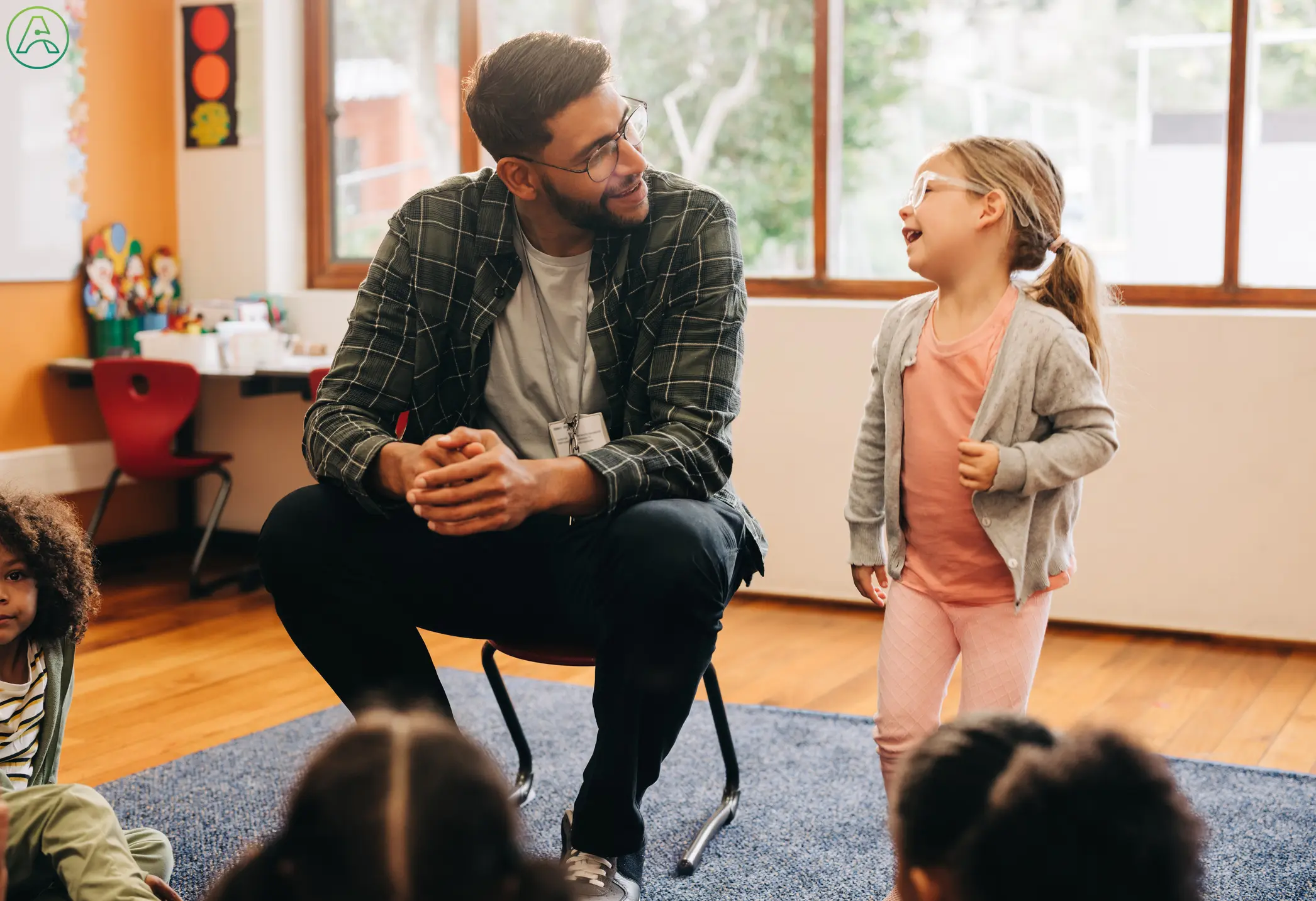 A little girl gives a speech to her preschool classmates while her teacher, a young man in a green flannel and jeans, encourages her with a smile.