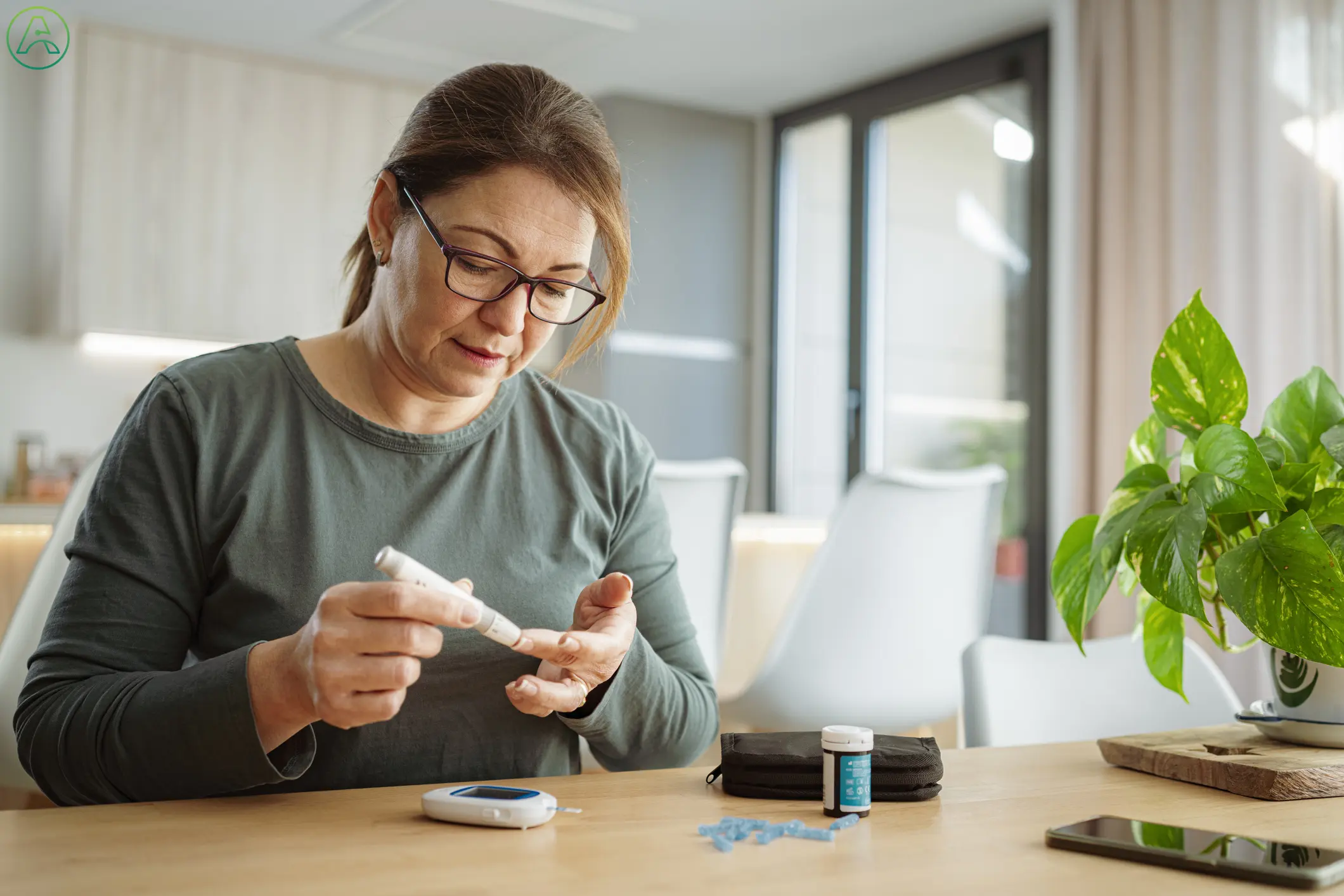 A mature white woman with reddish hair and black glasses uses a finger prick test to check her blood sugar levels while sitting at her kitchen table in a house decorated with white furniture and plants.