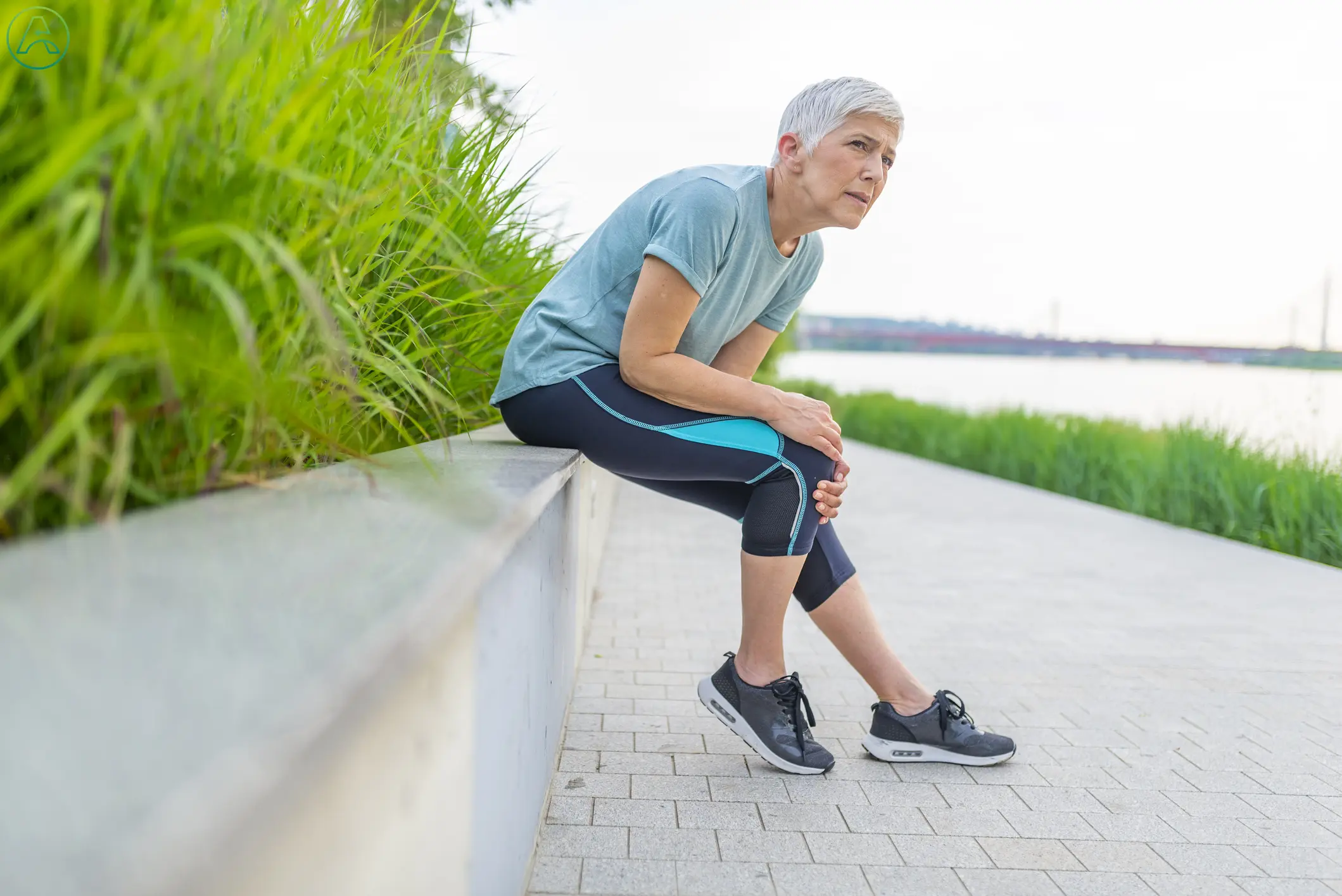 A middle aged white woman with short white hair and blue exercise clothes interrupts her run along a river to sit down and massage her knee.