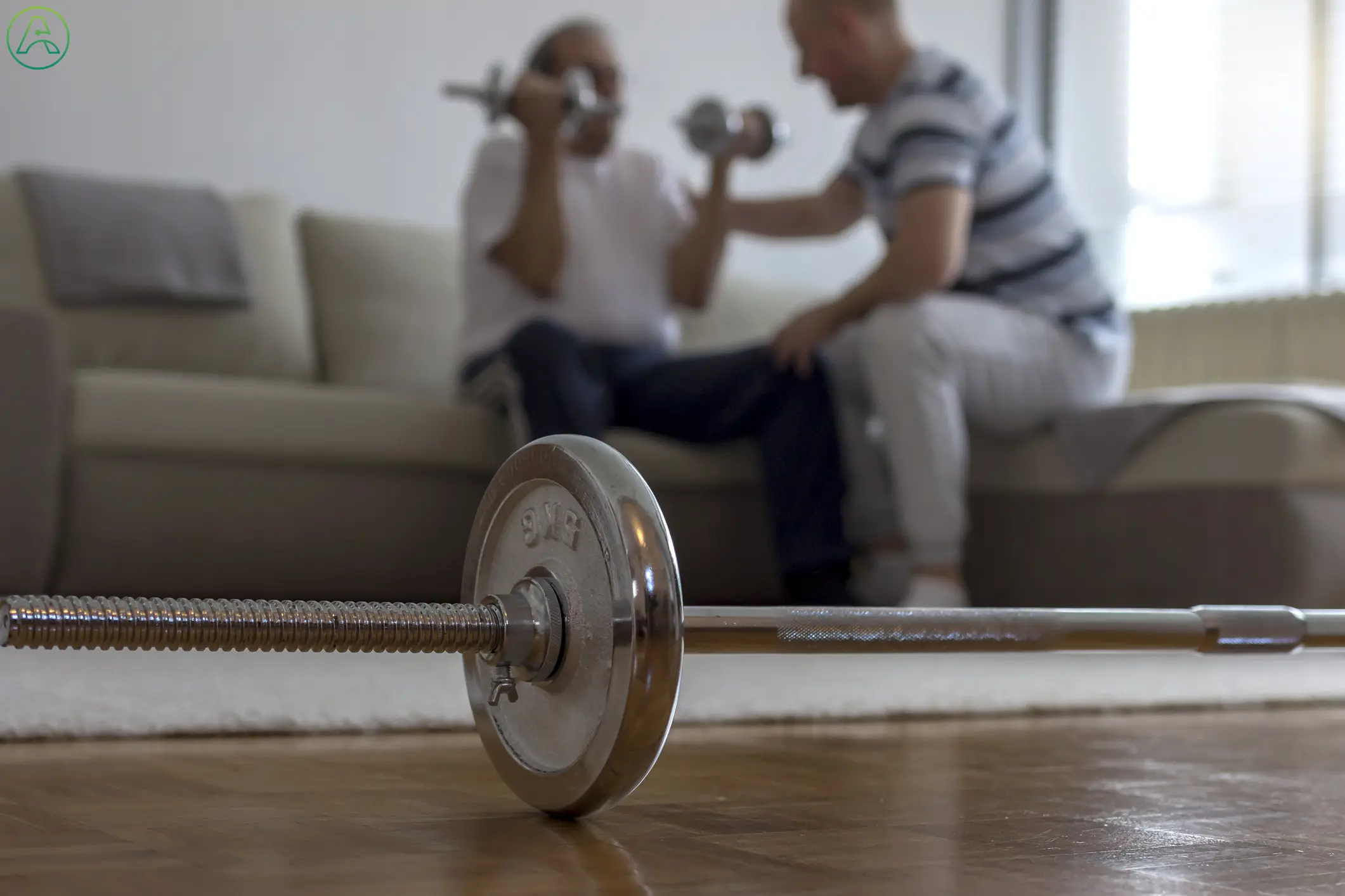 A physical therapist in white sweatpants helps an older man in loose workout clothes through arm strengthening exercises as he recovers from a stroke at home.