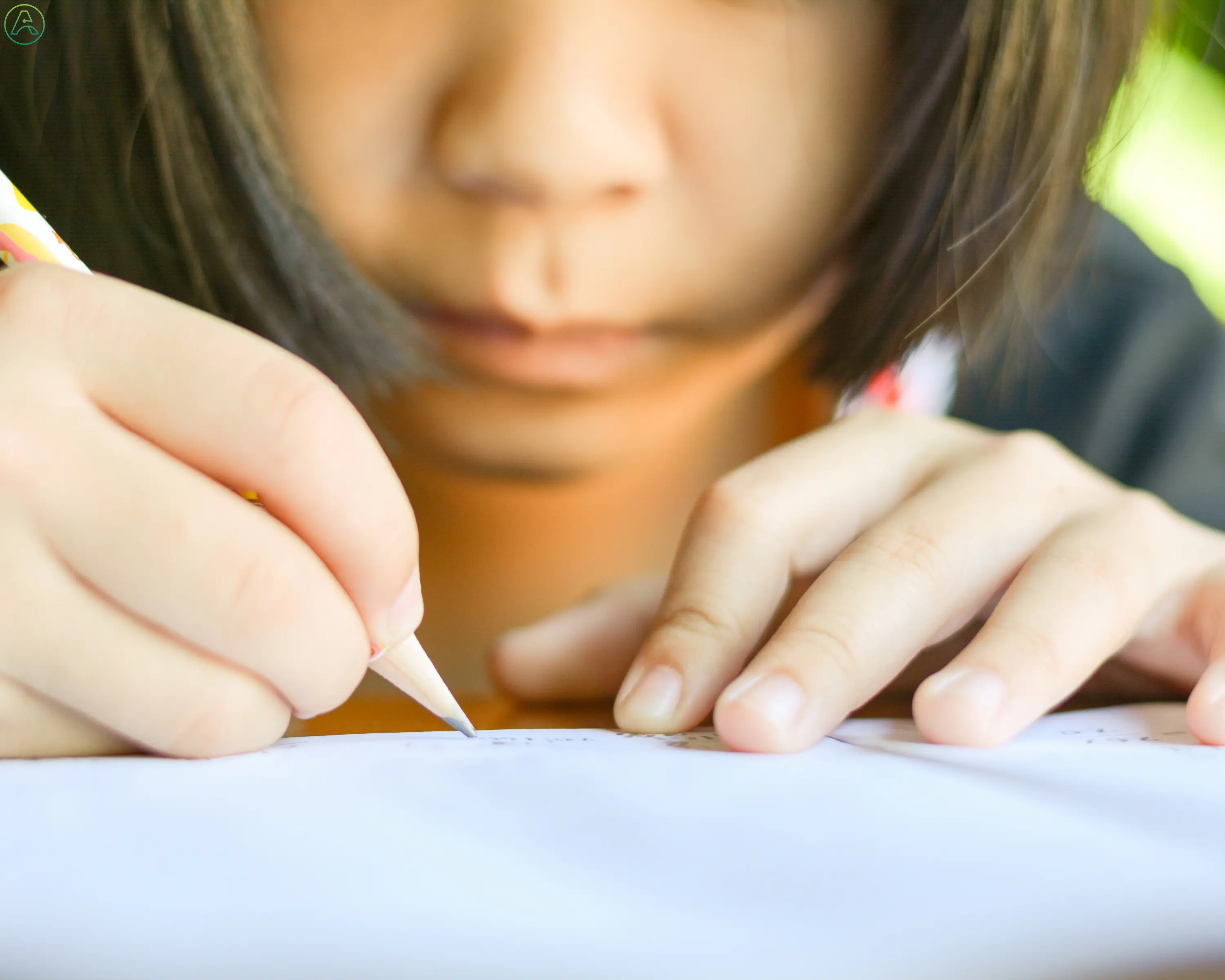 A schoolgirl with dark hair leans too close to her paper as she tries to write with a pencil she's holding awkwardly in her right hand.