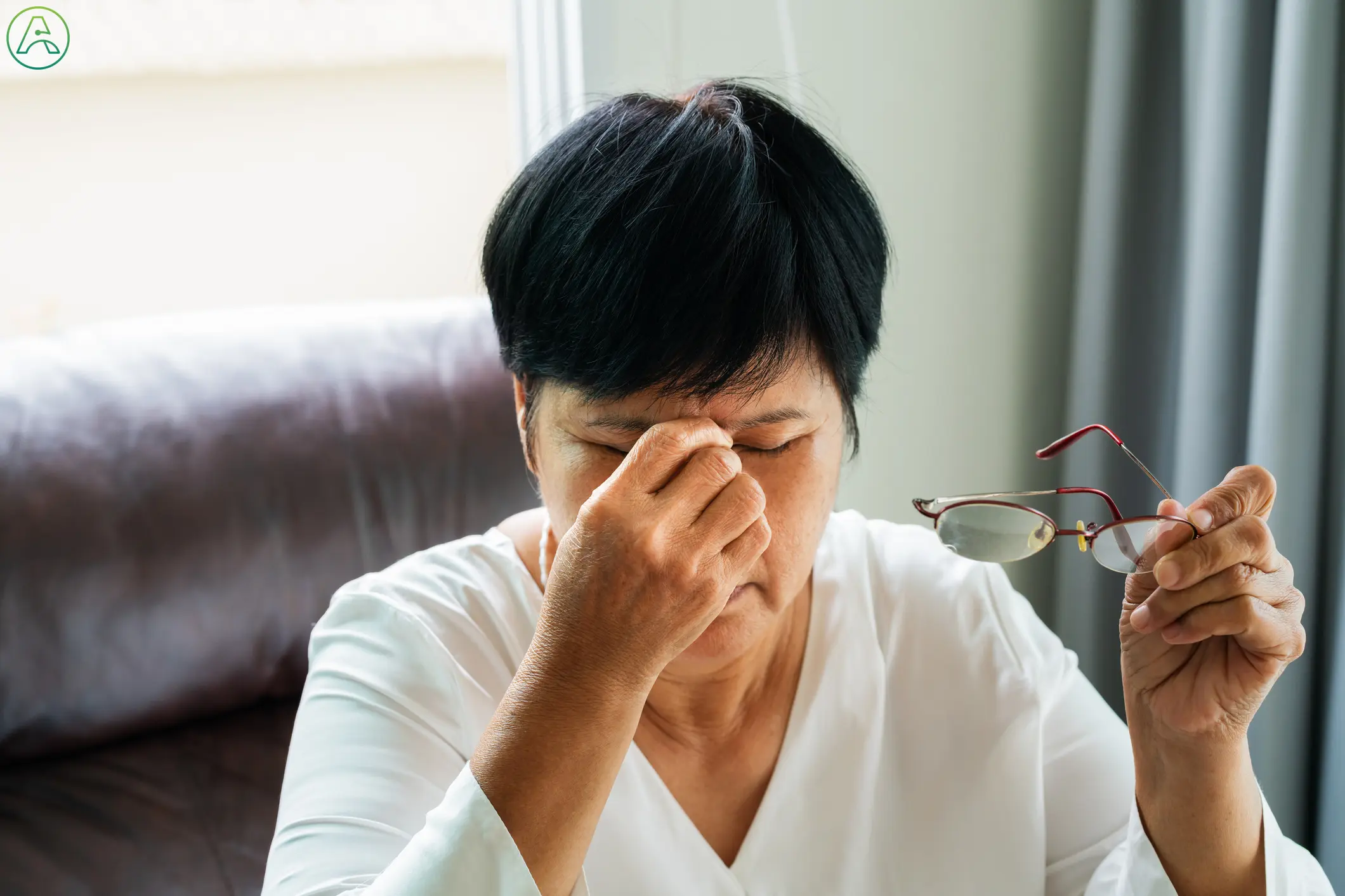 A senior Asian woman sits in her living room on the couch, rubbing her eyes in pain. She has removed her reading glasses and holds them in her hand.