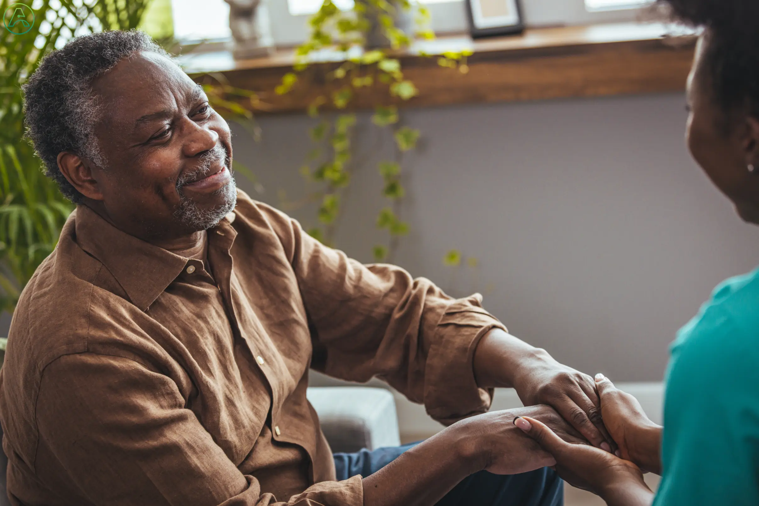 A senior Black man smiles at a Black nurse who holds his hands in support.