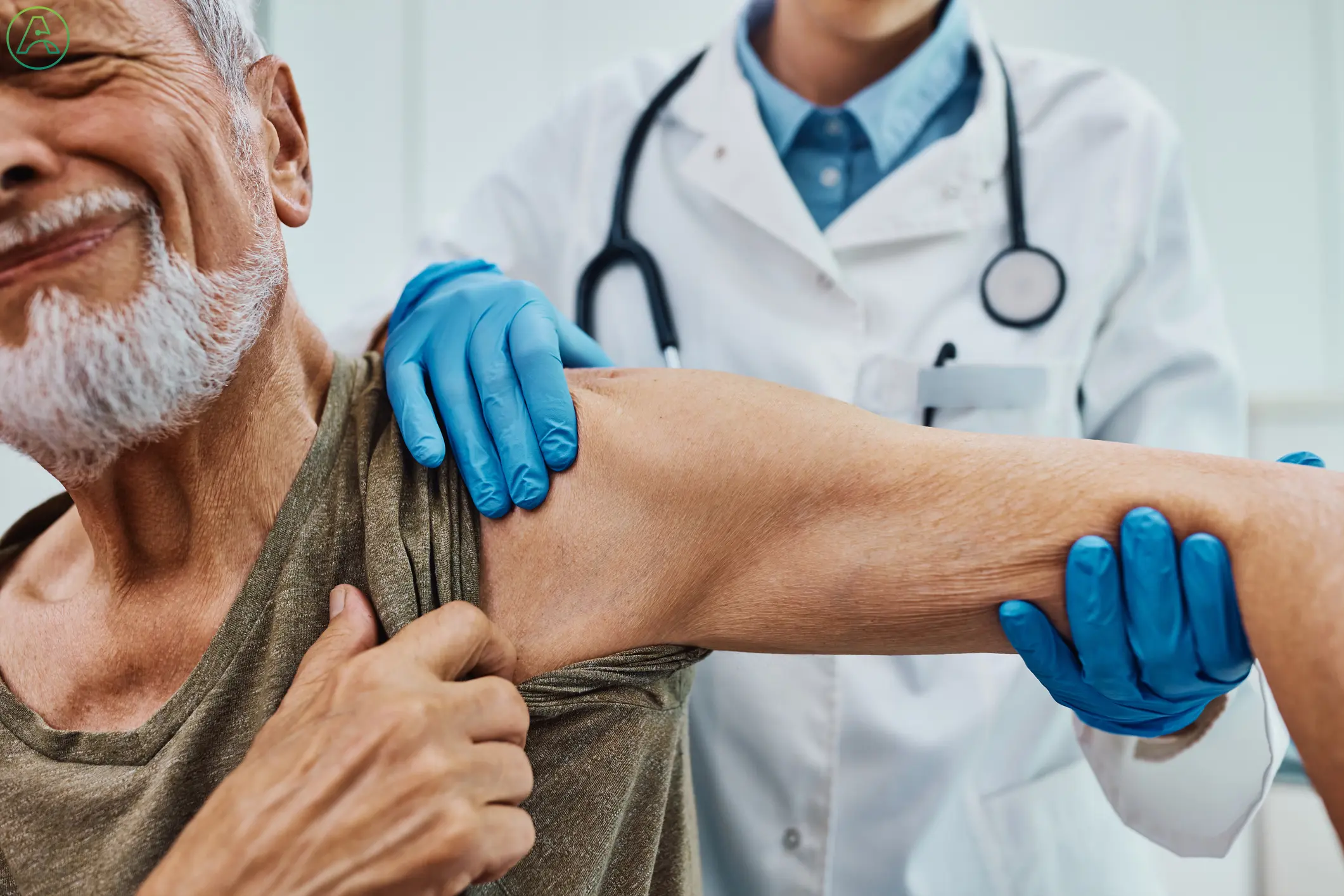 A senior white man in a dark green shirt grimaces in pain as a doctor in a white coat and blue gloves tests his shoulder joint for signs of arthritis.