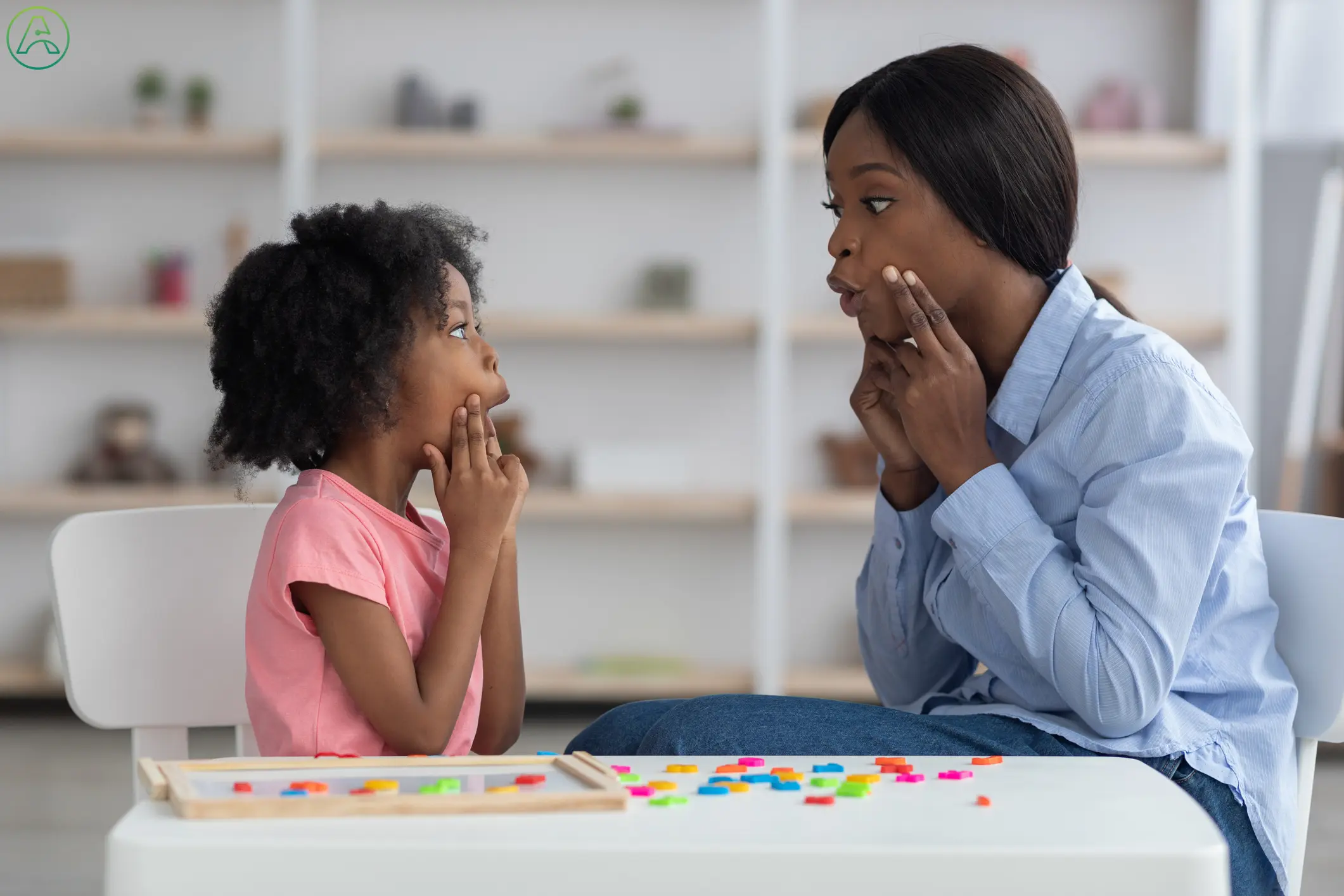 A speech language pathologist holds her fingers to her cheeks while her young female patient mimics the vocal exercise.