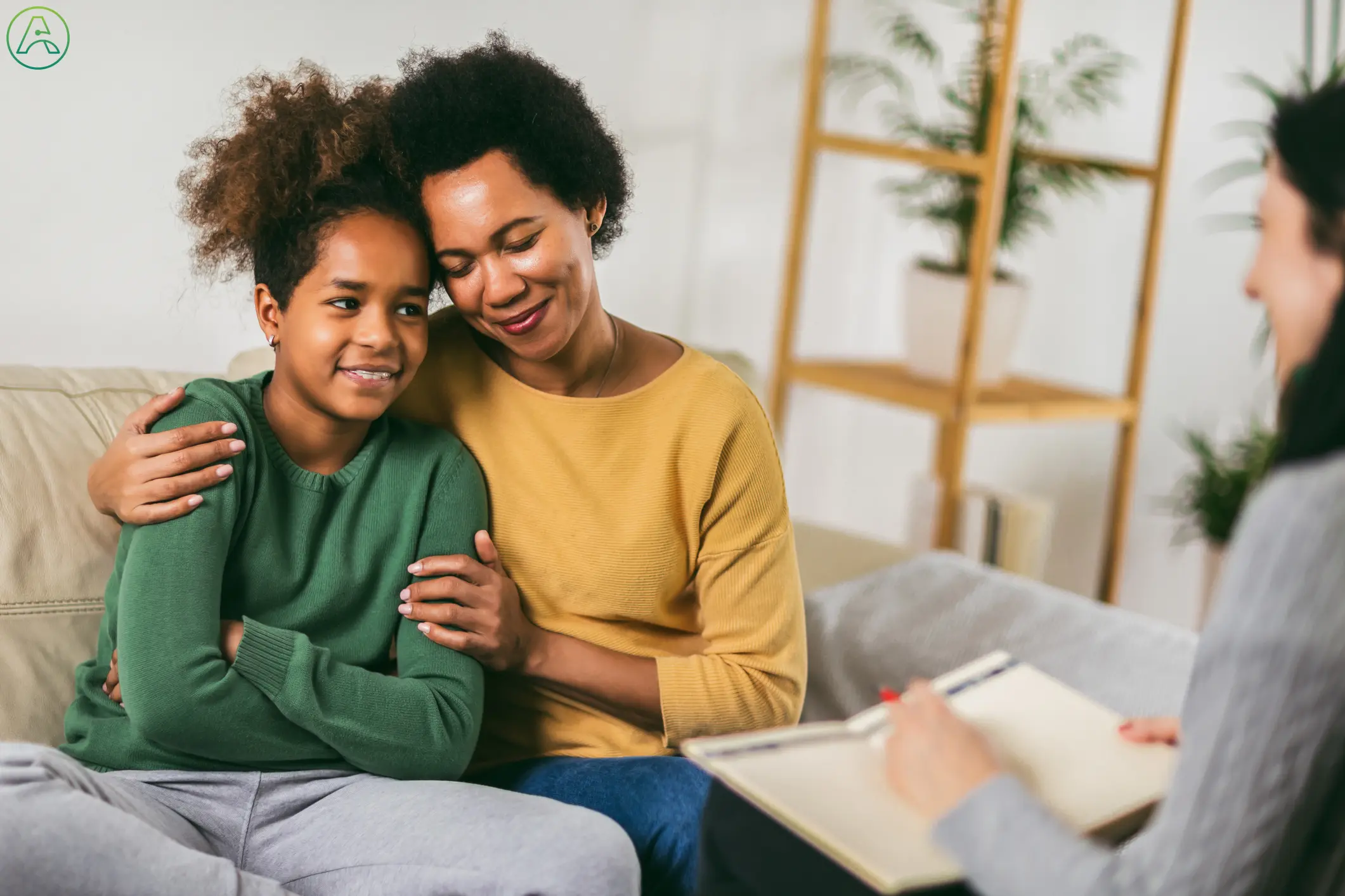 A teenage Black girl and her mother attend therapy together. The girl has her arms crossed tightly against her chest but is smiling.