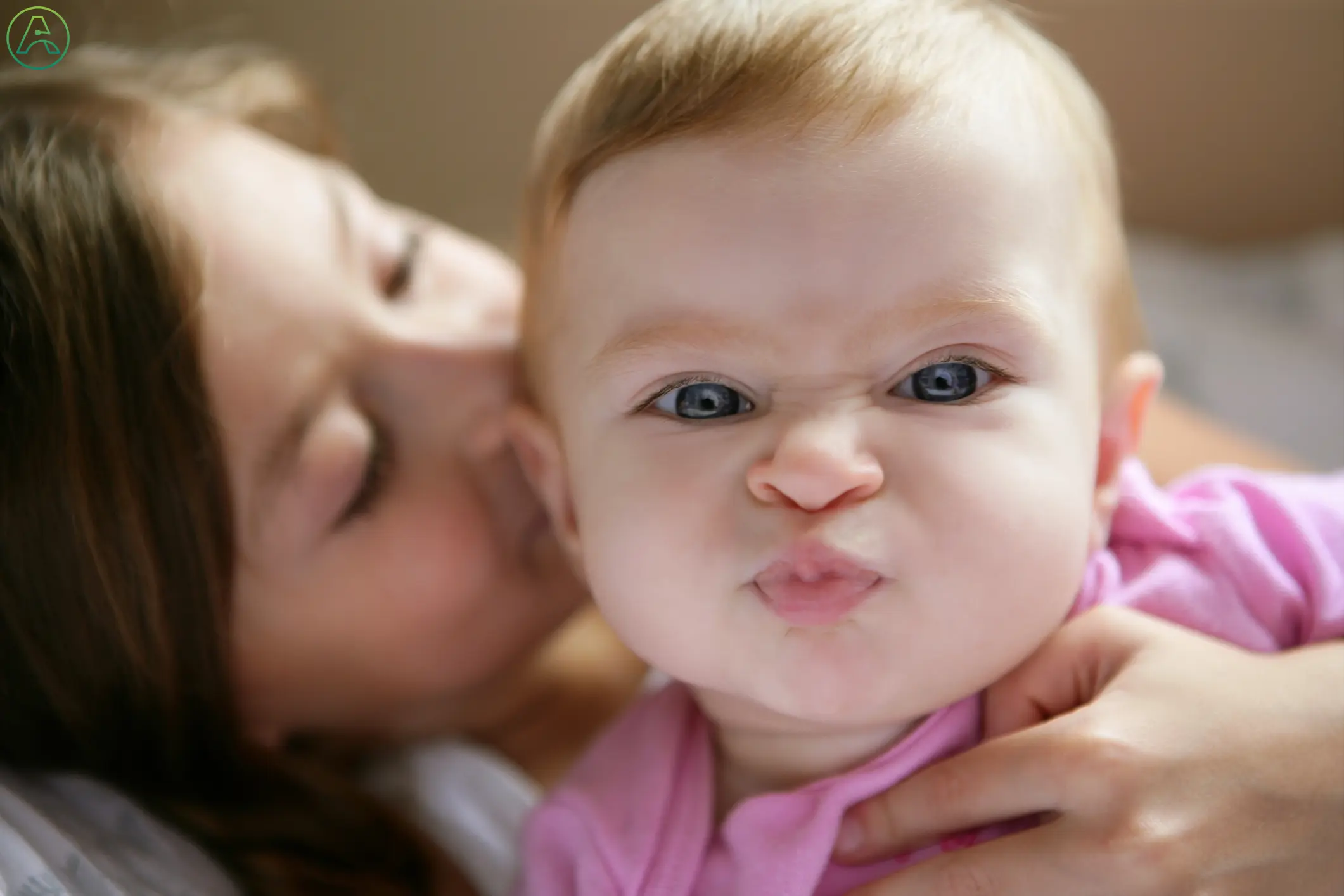 A white baby girl with red hair and blue eyes scrunches up her face at the camera while her mother kisses the side of her head.