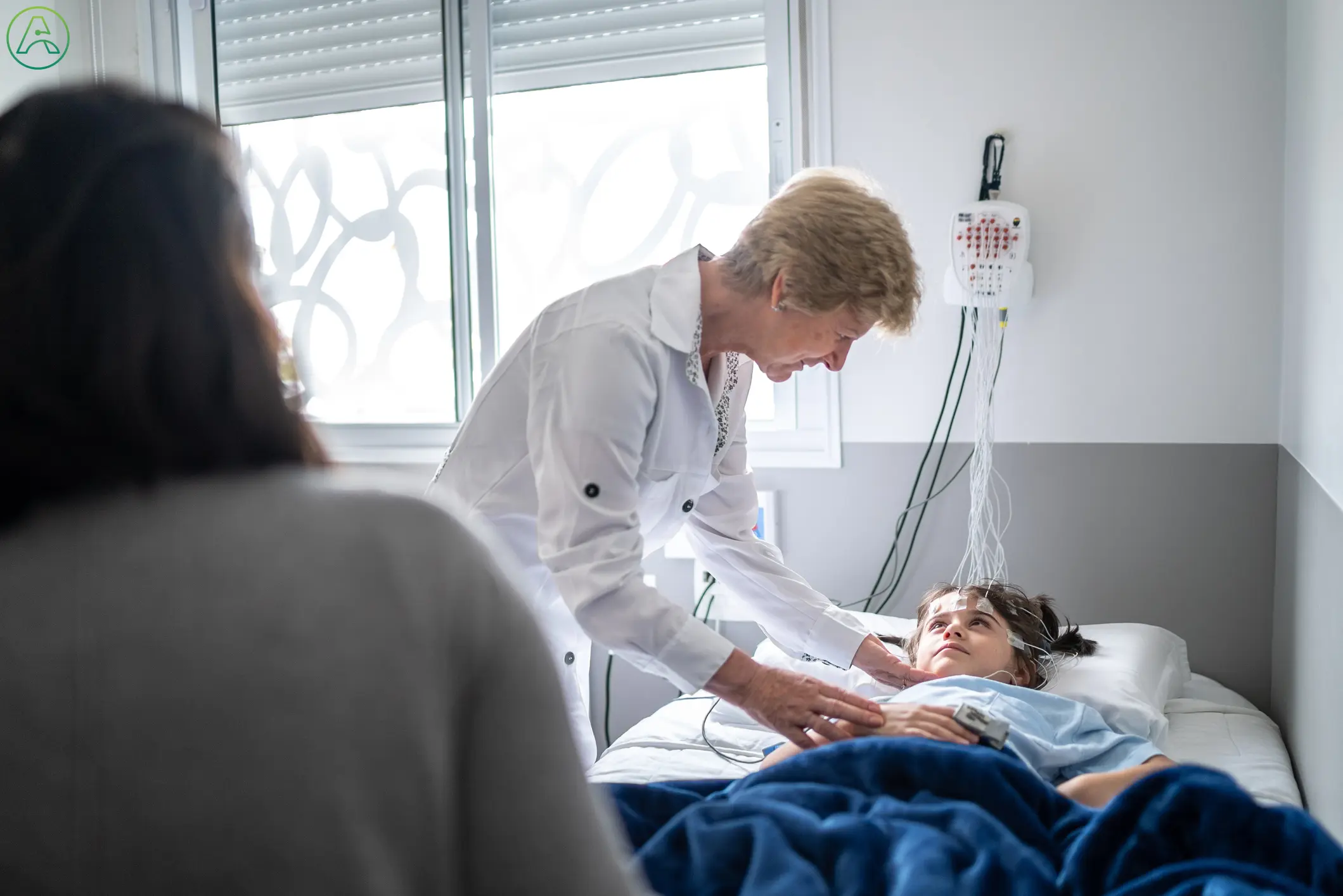 A white female doctor explains a sleep test to a young white child laying in a hospital bed with EEG electrodes attached to his head while his mother looks on.