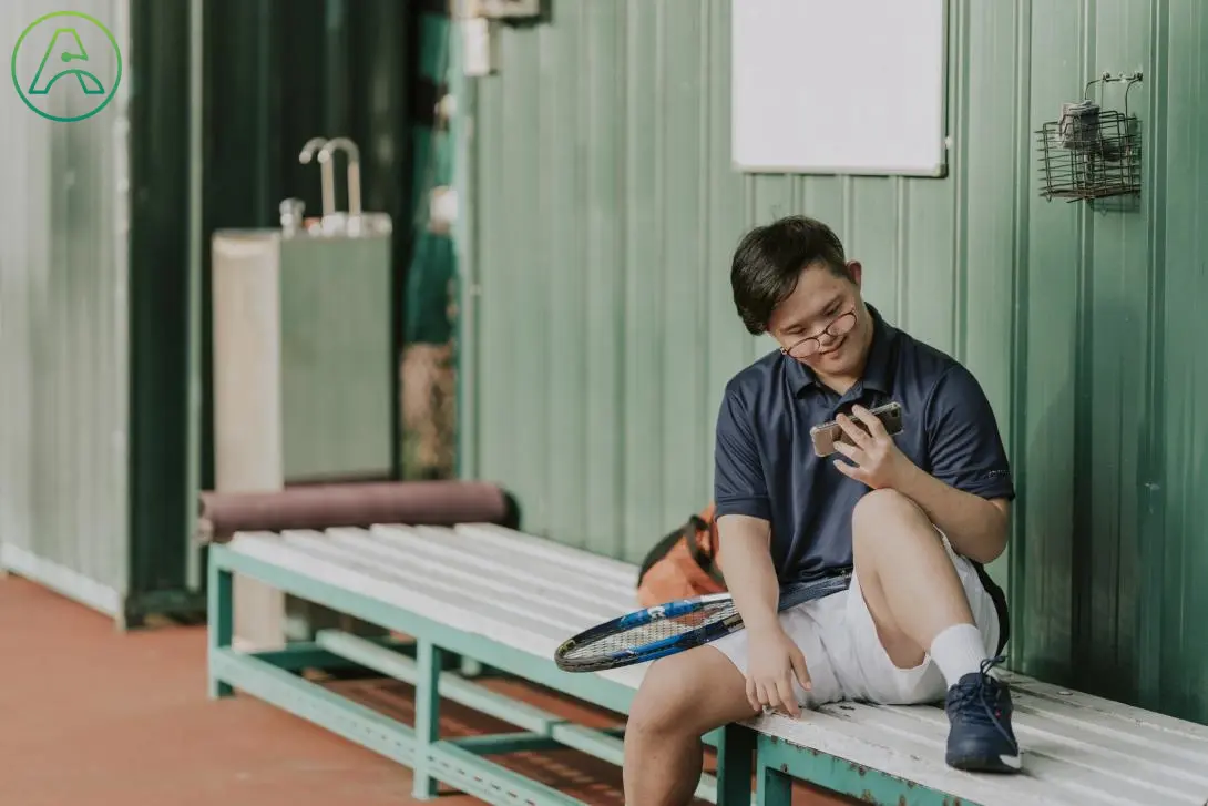 A young Asian man with Down syndrome wearing glasses and a collared black shirt takes a break from playing tennis to sit on a white bench in a green athlete's shade shelter, watching something on his phone.