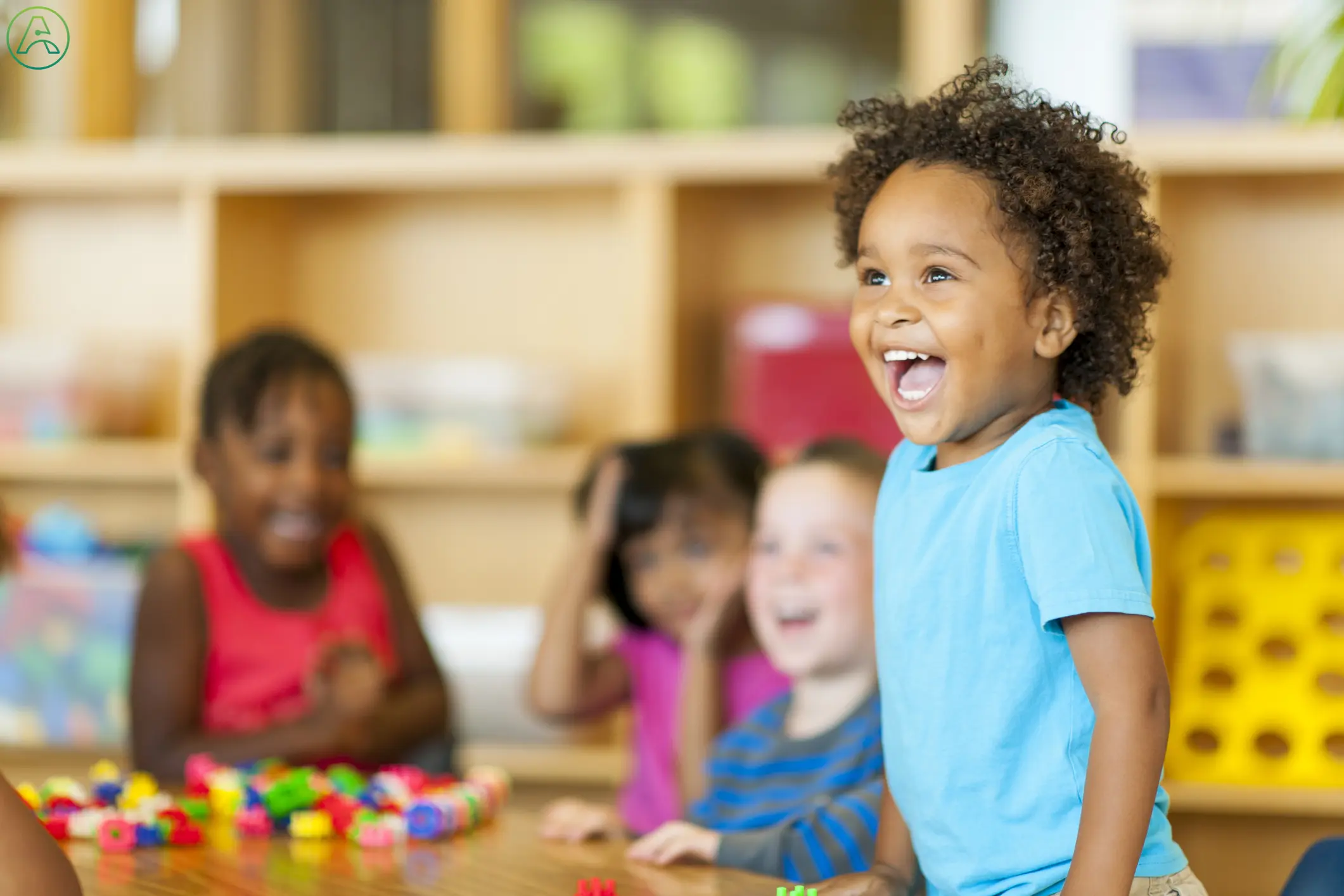 A young Black boy, having fun in his preschool classroom, laughs and leaps up from his chair while his other classmates stay seated behind him.