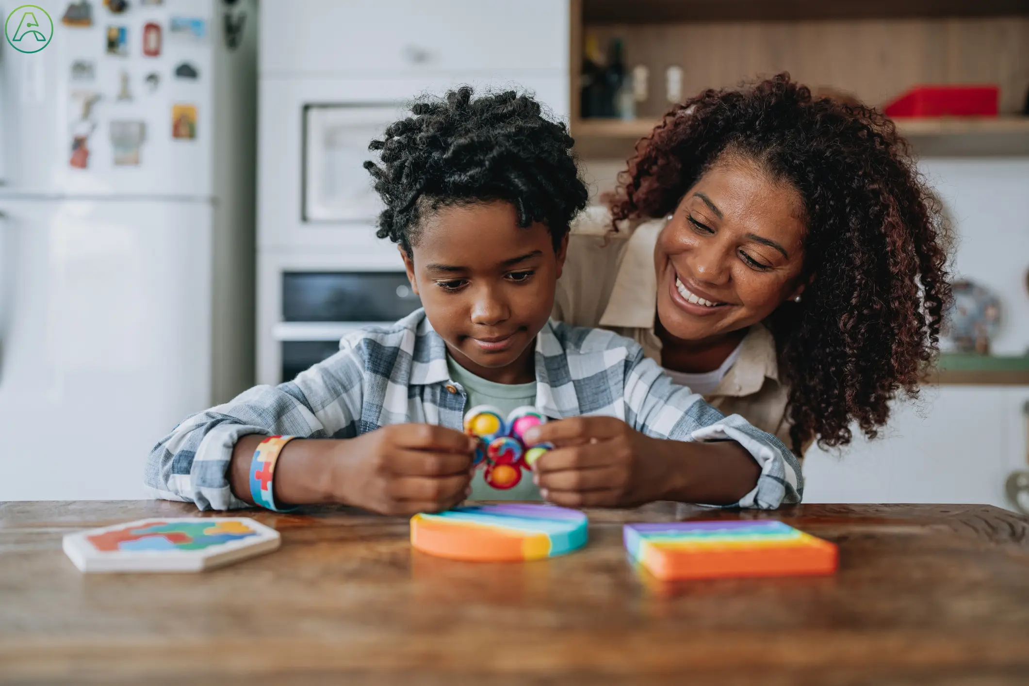 A young Black boy with curly black hair and  a gray plaid button-down shirt sits at a kitchen table playing with fidget toys while his smiling mother watches from behind him.