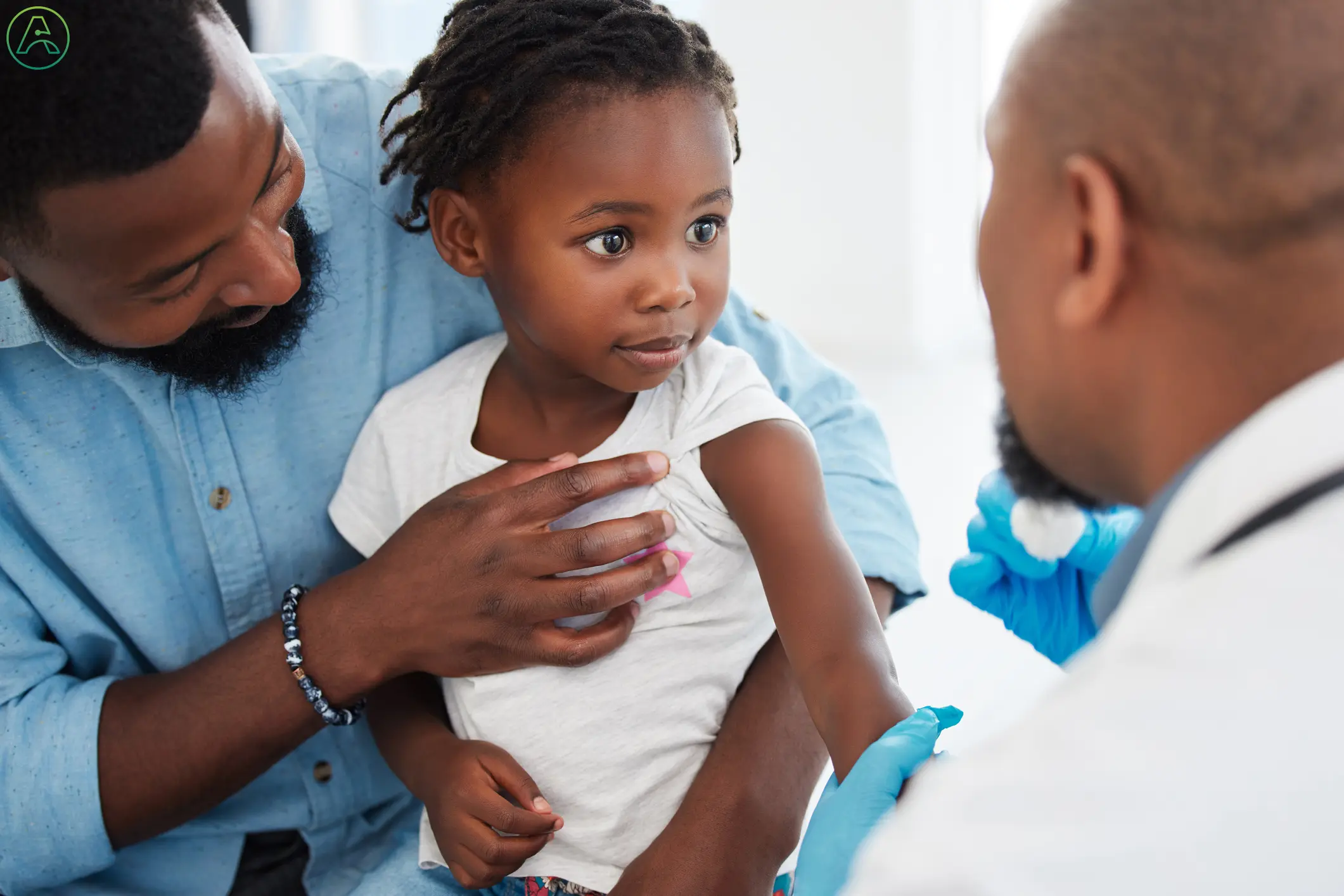 A young Black father protectively holding his toddler daughter while she looks up at a doctor with a nervous expression.