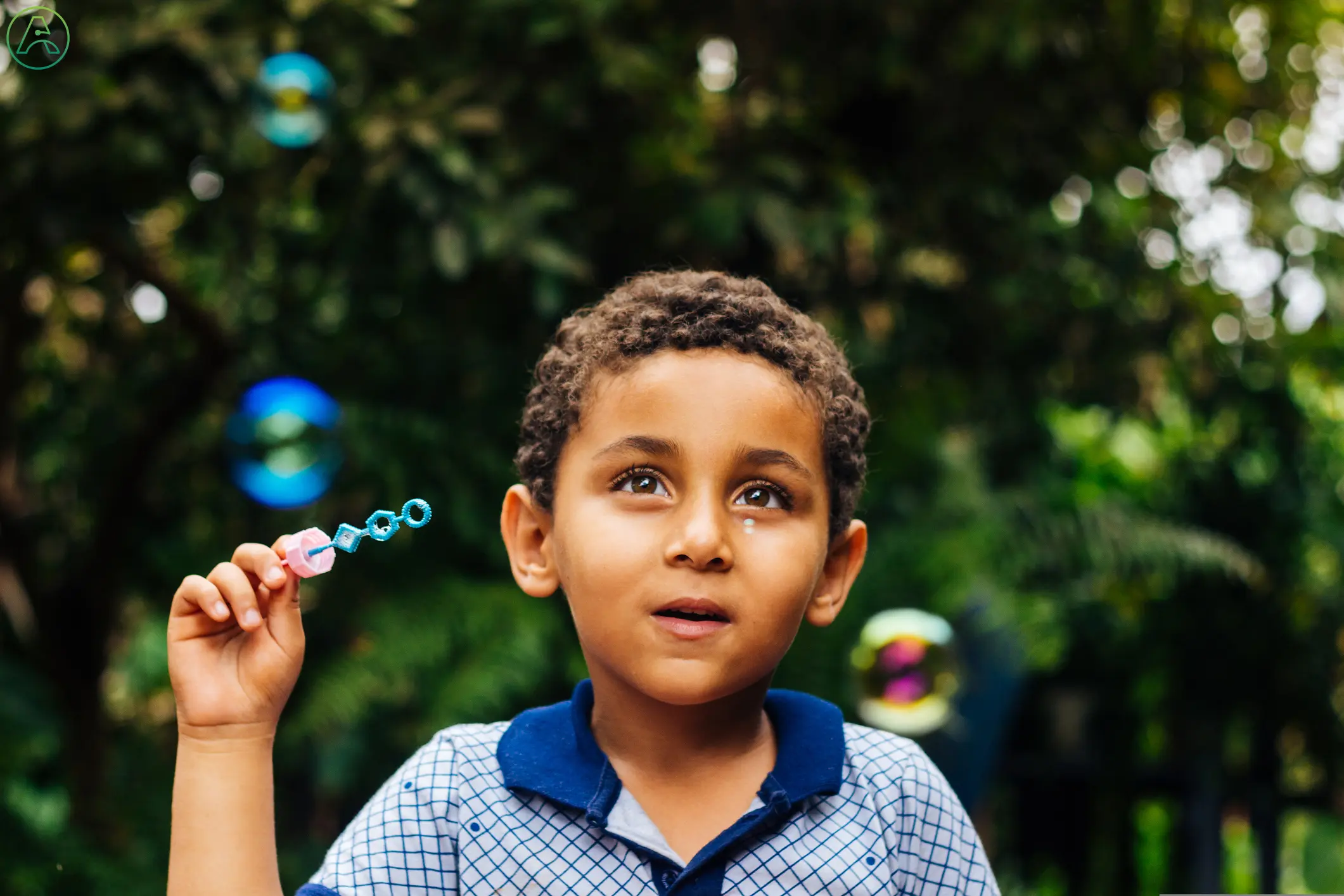 A young brown-skinned boy squints at rainbow-colored bubbles against a green leafy background.