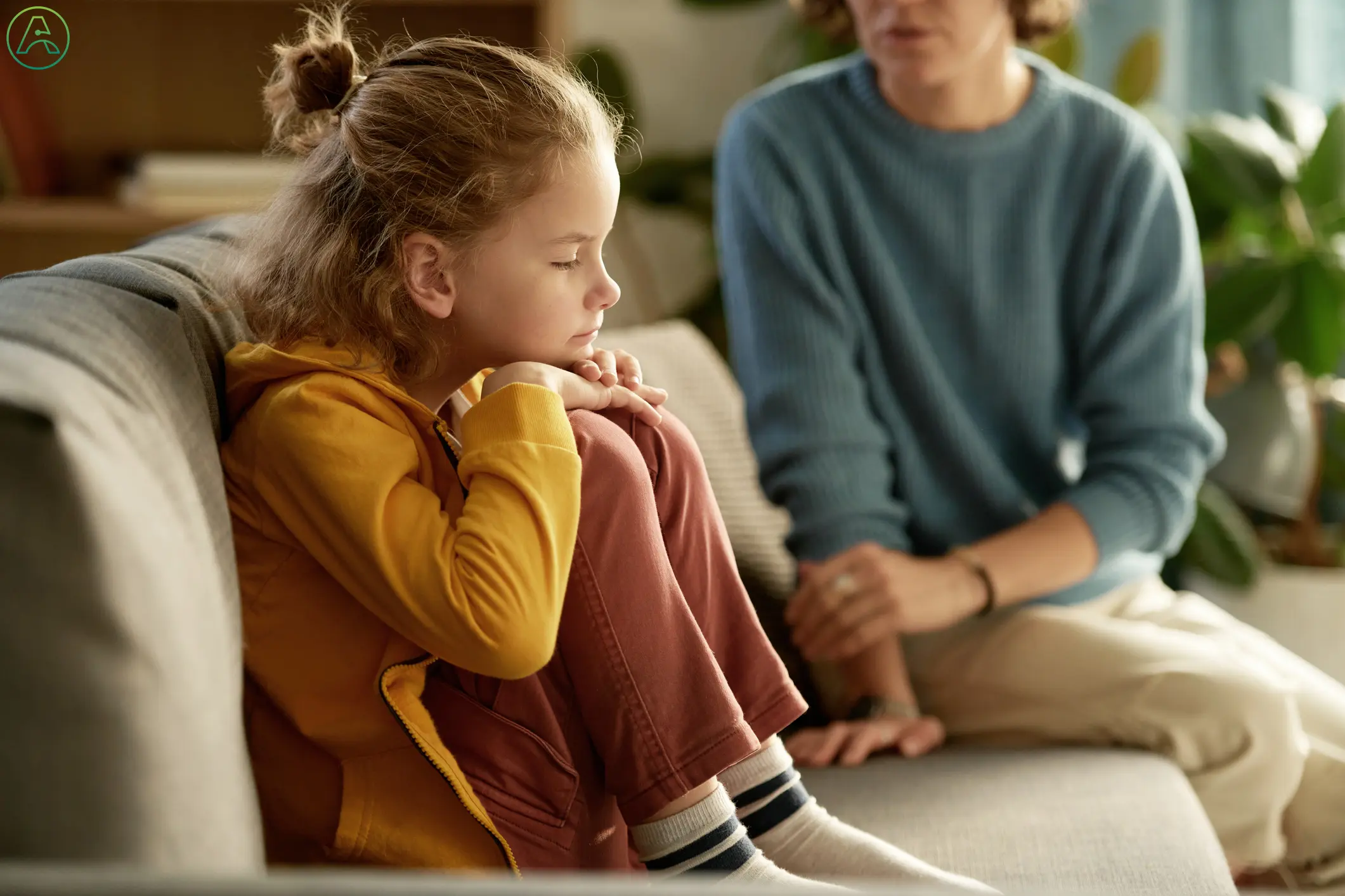 A young child in a yellow sweatshirt and red pants curled up tight on a couch, unable to speak to the concerned mother sitting beside them.