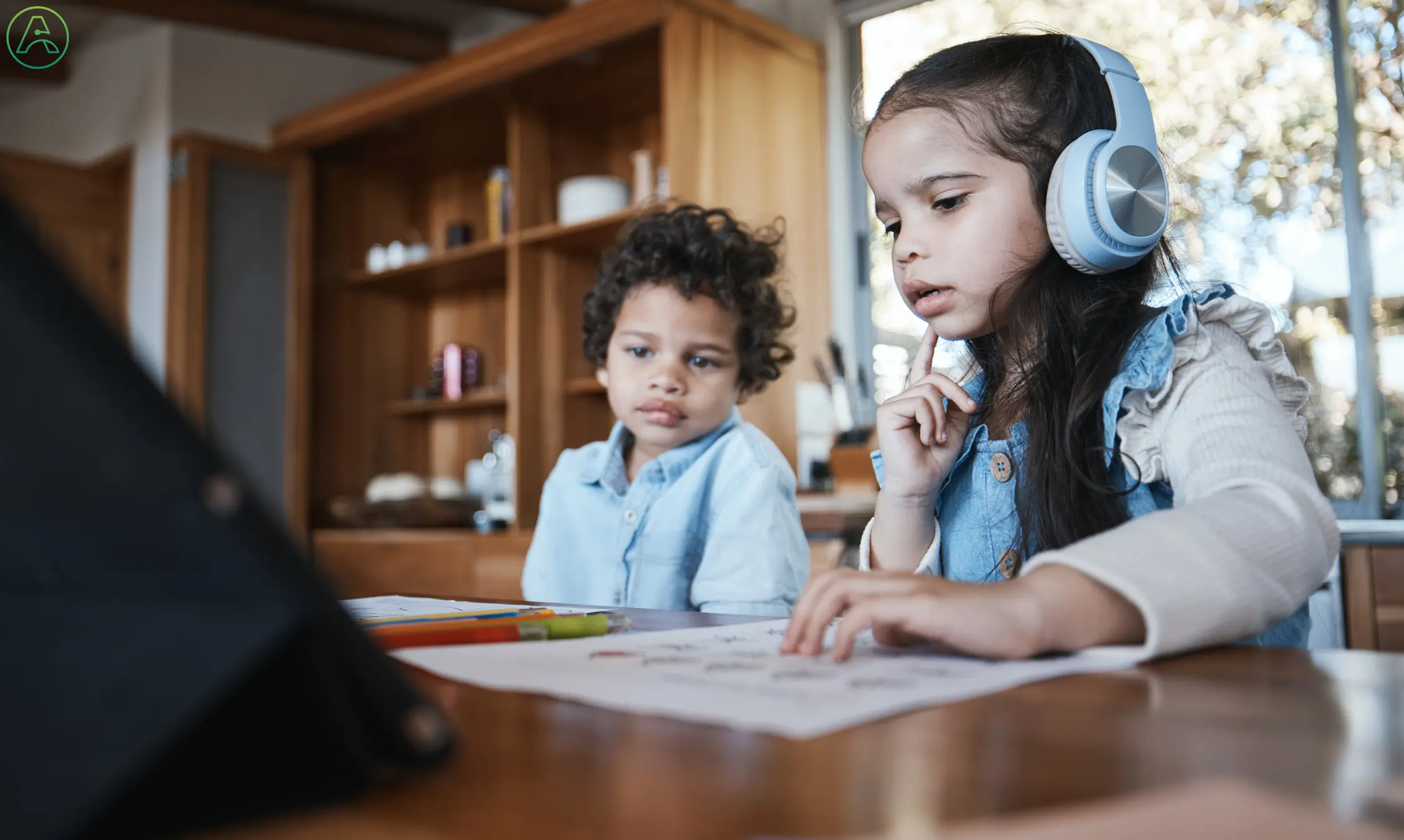 A young girl with wavy brown hair wears noise-cancelling headphones so she can focus on her homework while her younger brother tries to distract her.