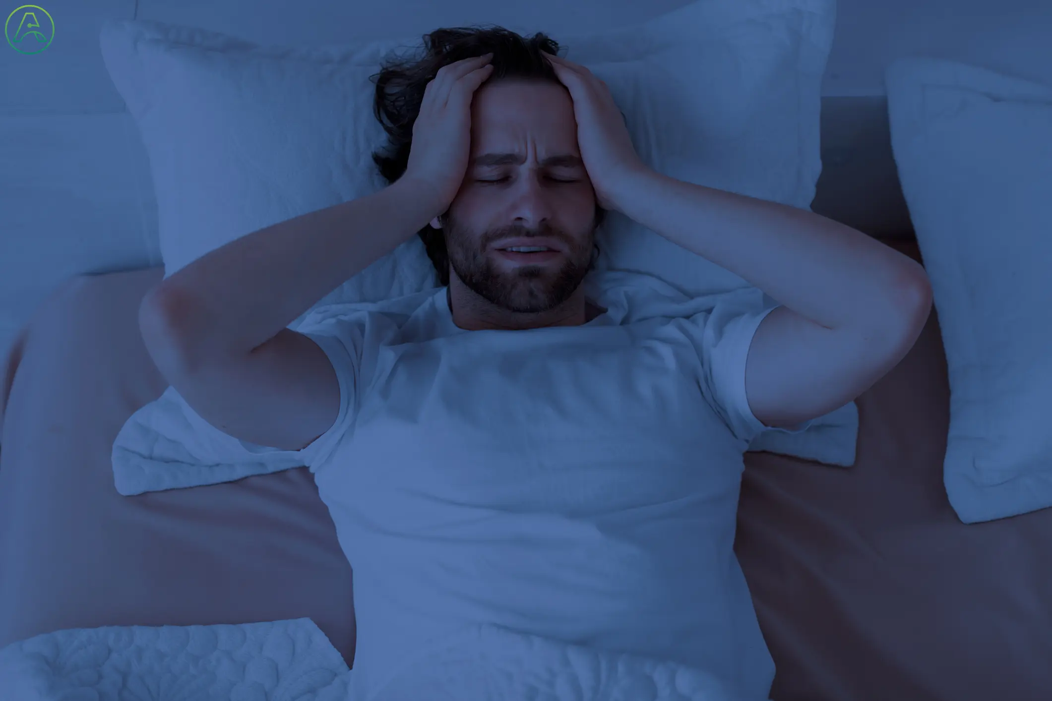 A young white man lays in bed before dawn, holding his head in both hands in frustration at not being able to sleep.