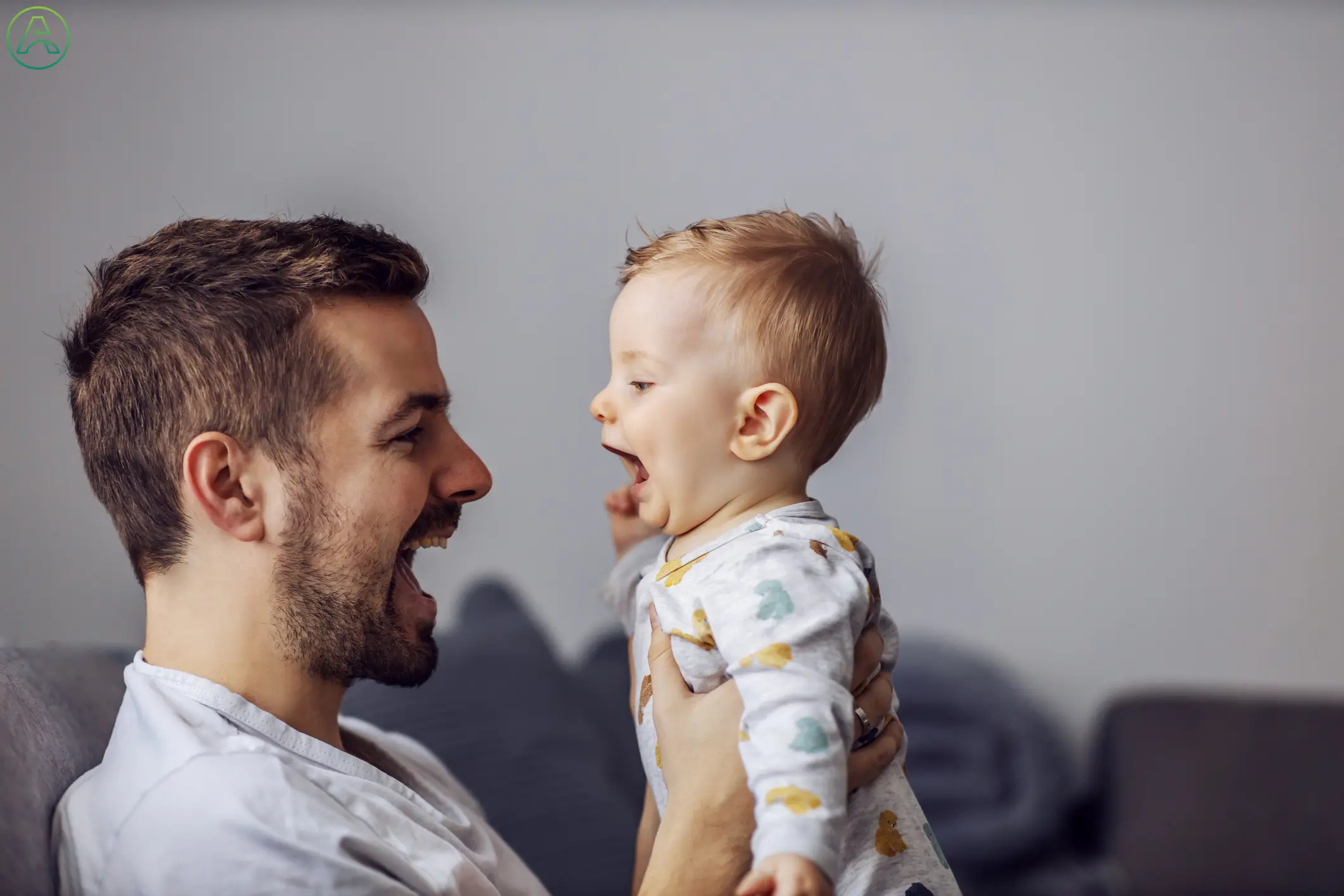 A young white father and his toddler son play on the couch together at home. The baby, wearing a leaf-patterned onesie, repeats the noises his dad makes.