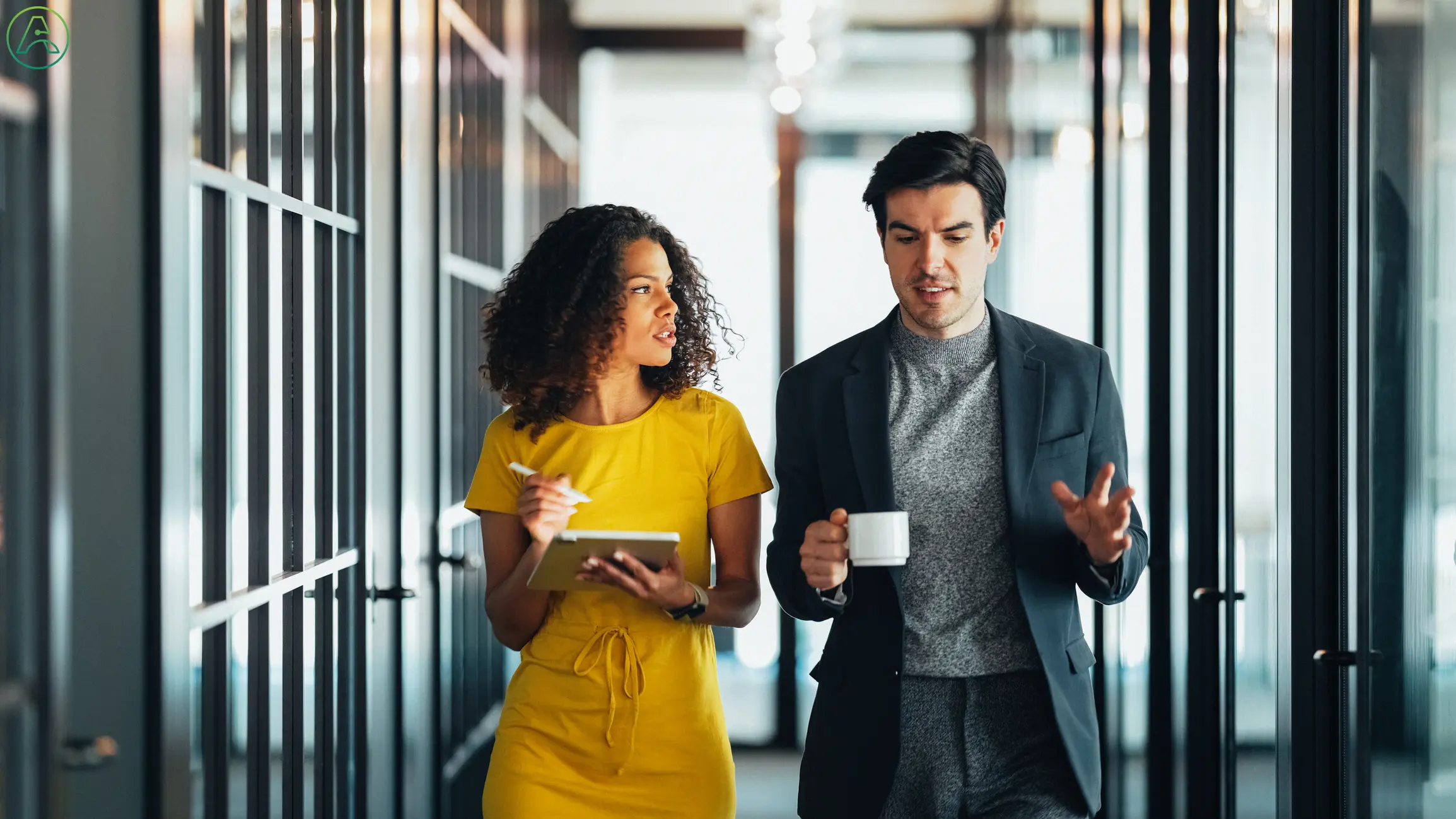 A young white man wearing a black business suit and a grey turtleneck lays out his day's goals in the hallway of a modern coworking space while his boss, a mixed-race woman with long curly hair and a yellow dress, helps him prioritize.