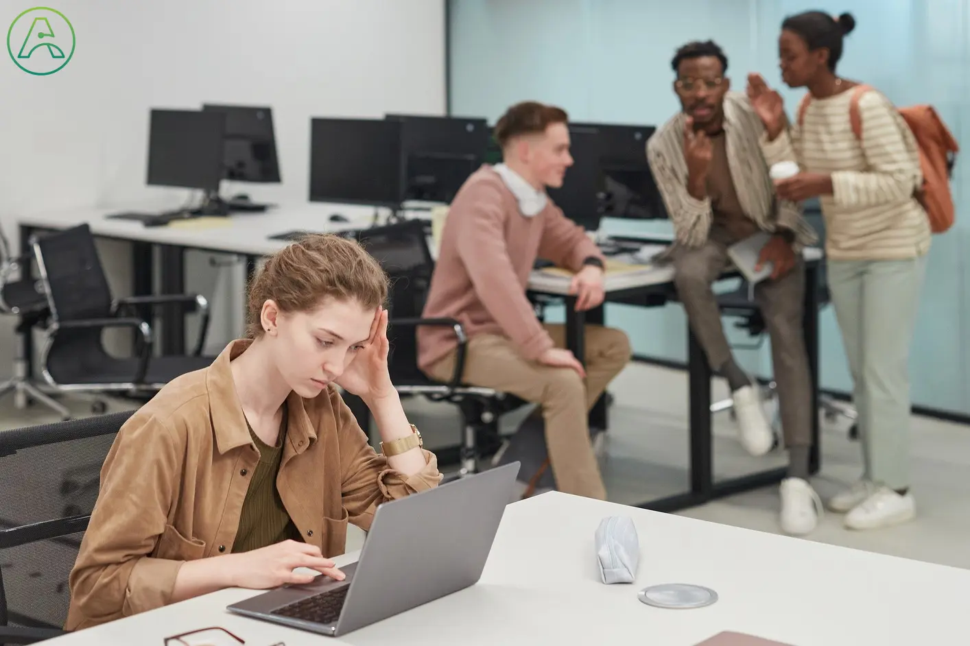 A young woman in a brown button down shirt sits in a technology startup, working alone because she can't contribute to her coworkers' conversation.