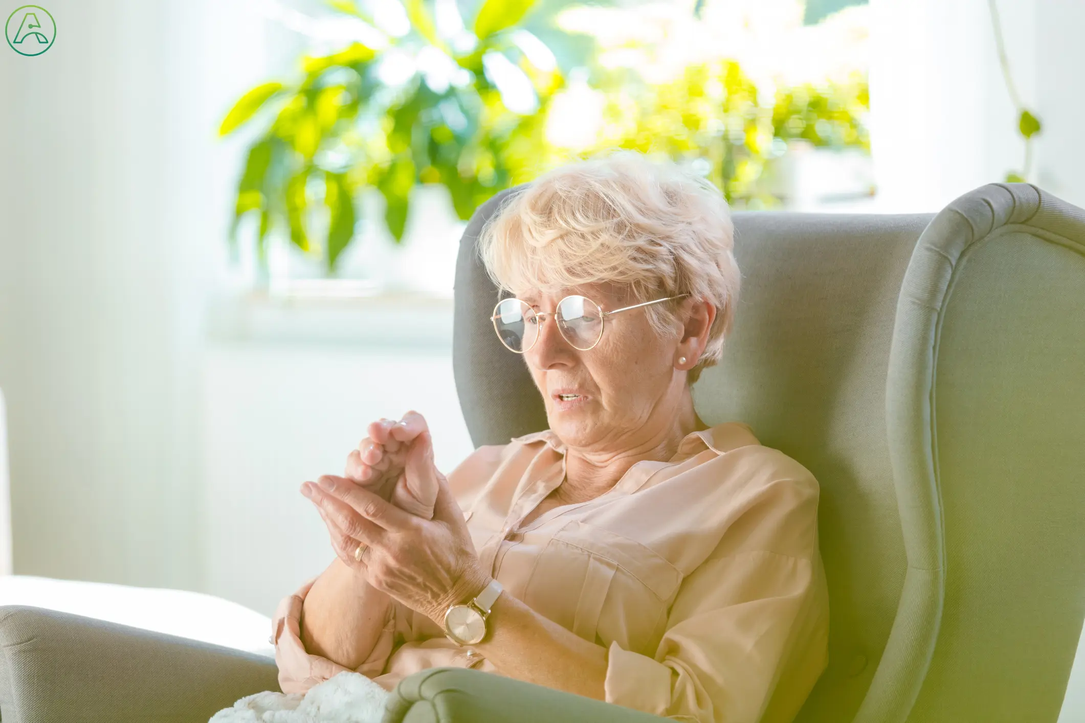 An elderly white woman wearing wire framed glasses and a light pink blouse sits in a green armchair at home, worriedly holding her shaking hand as it forms a claw shape.