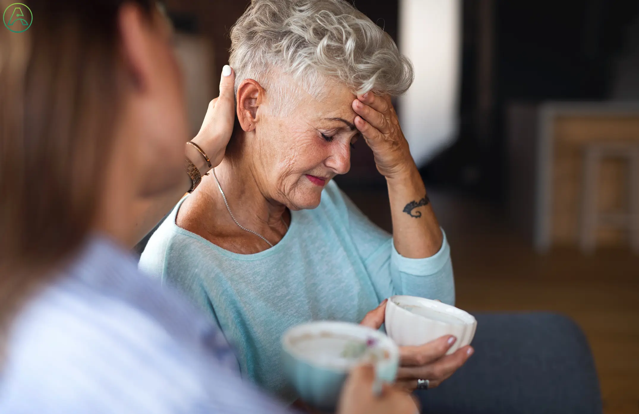 An older white woman struggles to hold a cup of coffee while her daughter reaches out to help.