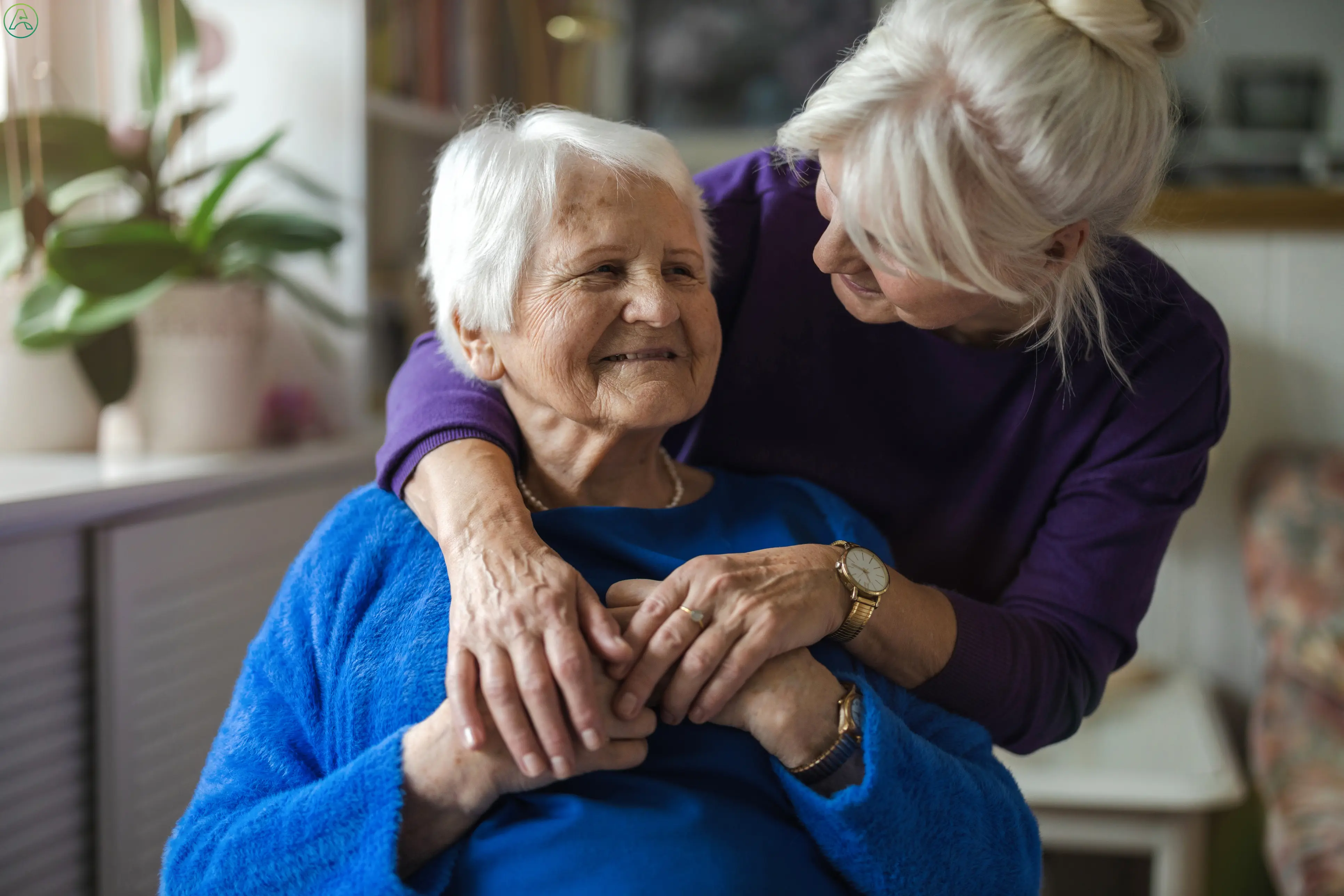 An older white woman with gray hair hugs her white-haired mother from behind.