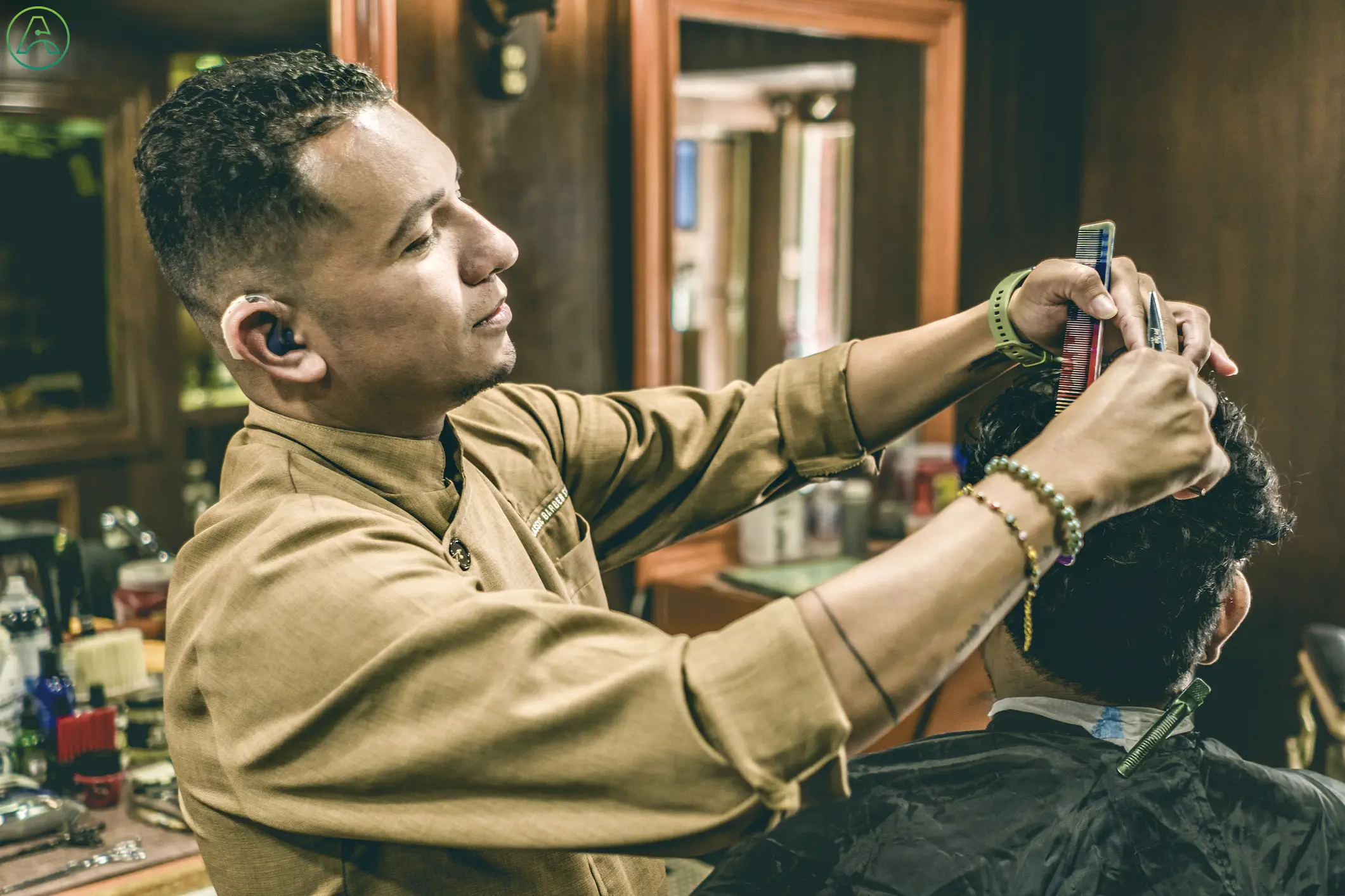 A barber with a pressed tan shirt and hearing aids smiles while talking with his client as he cuts their hair in a modern barber shop.