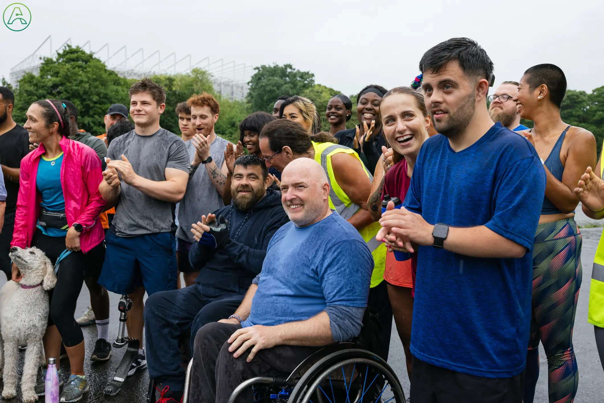 A diverse group of people, including individuals with disabilities, are gathered outdoors in an inclusive community event. The scene features people of different ages and ethnicities smiling, clapping, and socializing. Two men in wheelchairs are prominently featured in the center, smiling and engaged with the group. One man is wearing a prosthetic leg, and there are various participants in athletic wear, suggesting a sports or charity event. The atmosphere is positive, supportive, and celebratory.