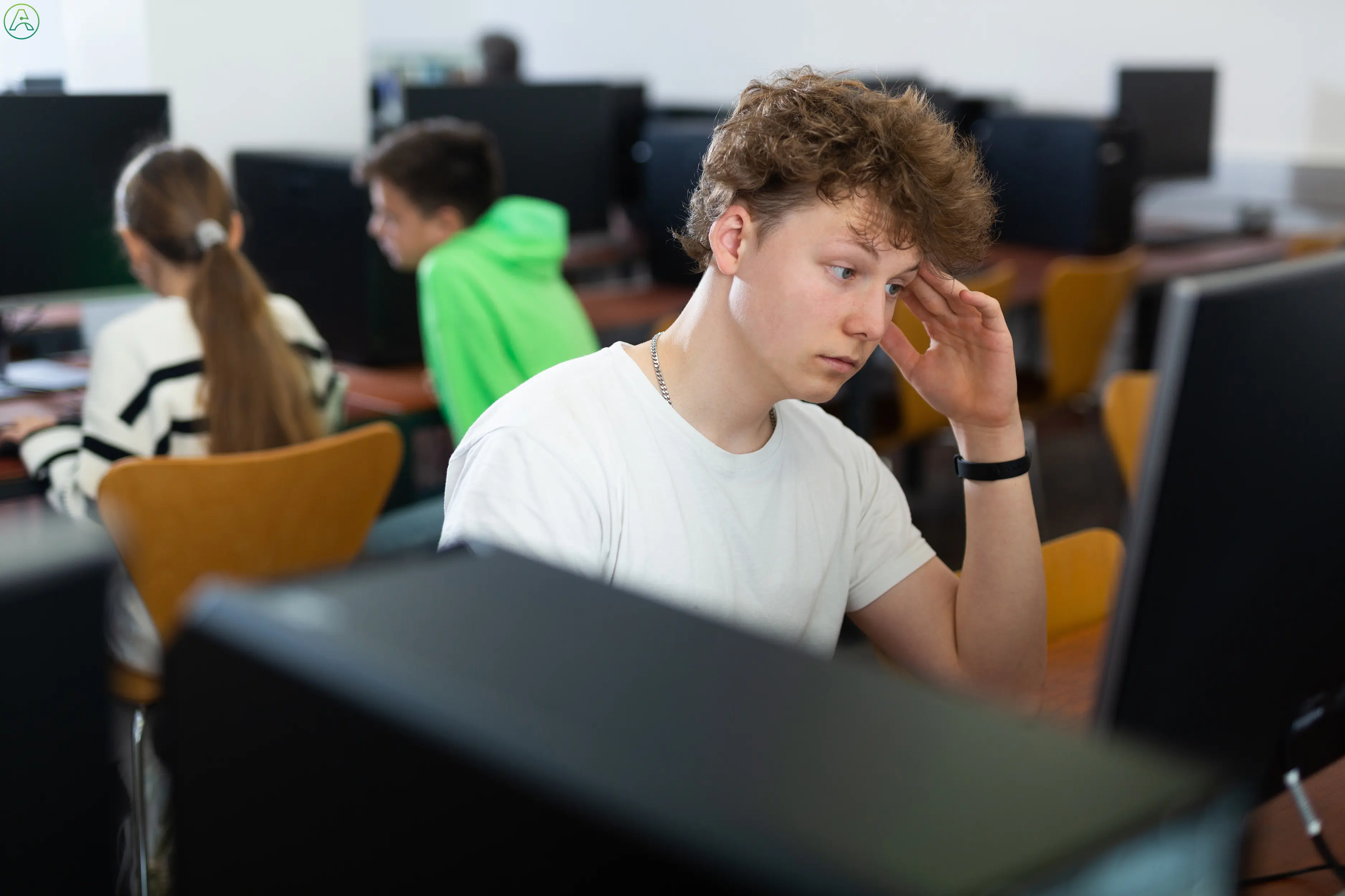 In a college computer lab, a frustrated student in a white tee shirt strains to drown out other people's conversation and focus on his assignment.