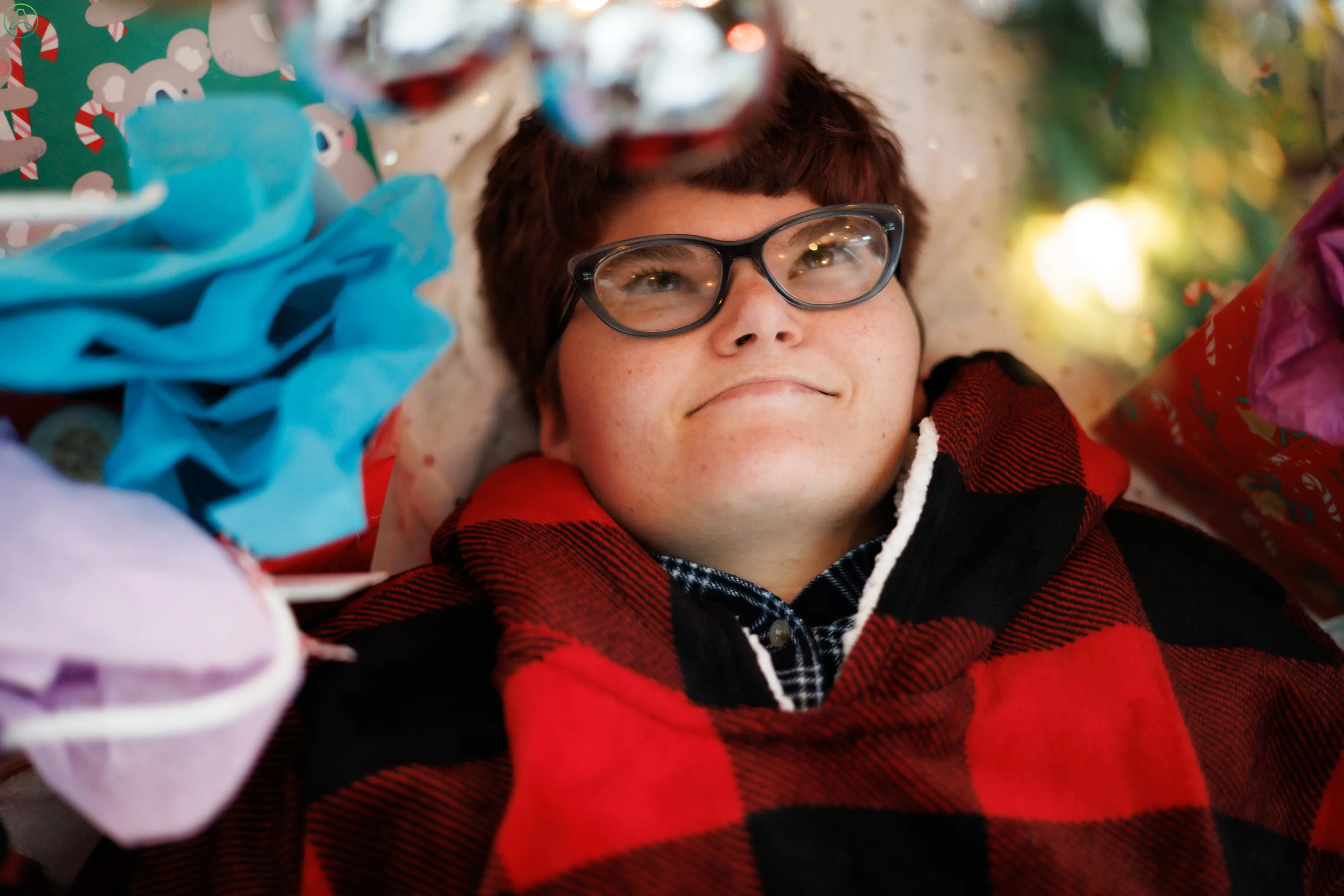 A young, white, red-haired person with an intellectual disability wears gray glasses and lays on the floor amidst a holiday mess of gift bags, lights, and decorations. They look up and smile softly, taking a break from the stress.