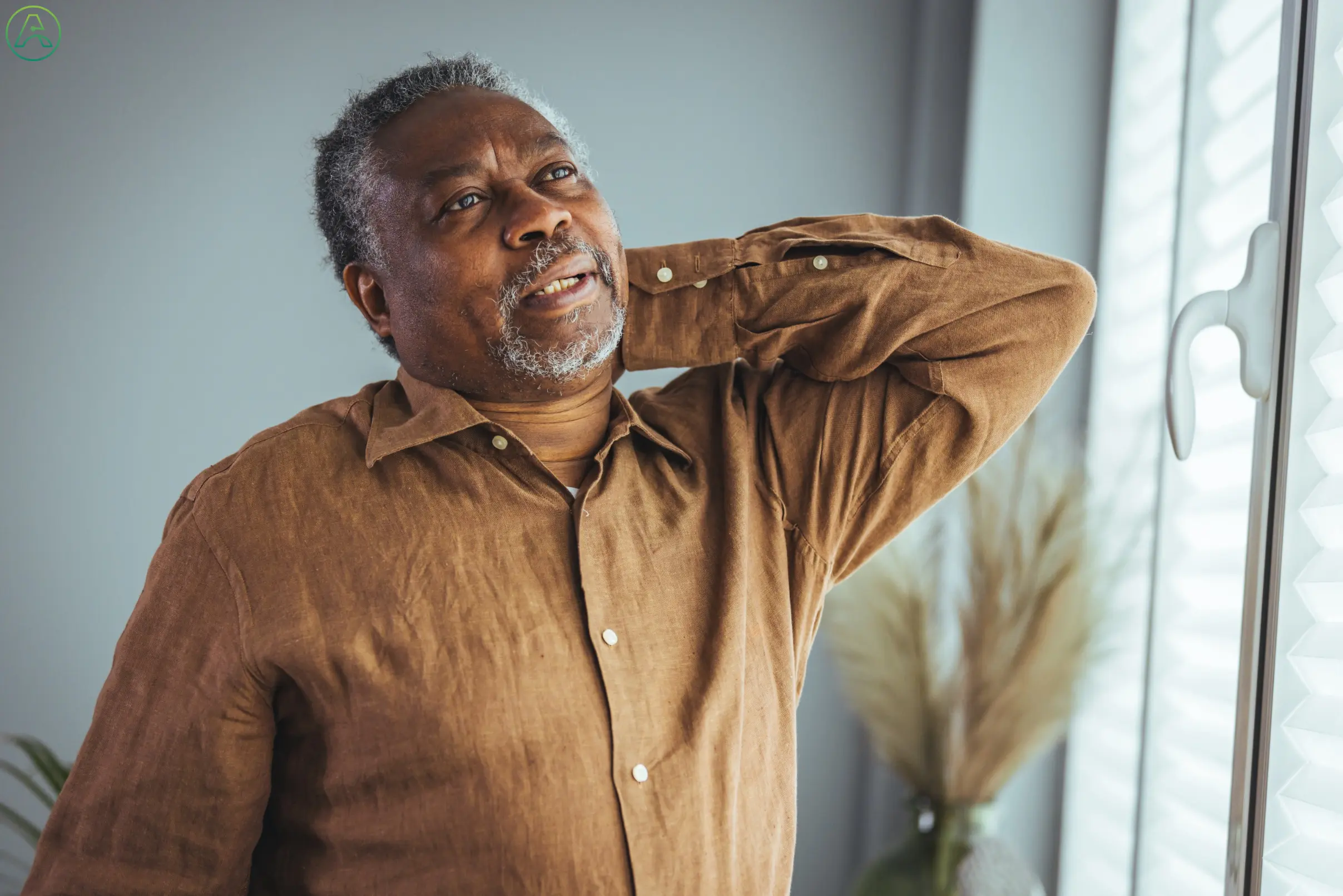 Senior Black man looks out window, rubbing back in pain.