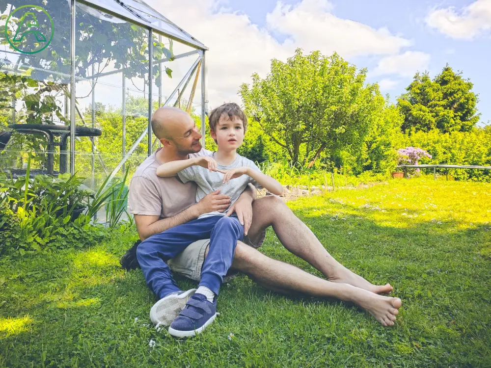 Sitting outside in the grass in front of a greenhouse, a young white father with a shaved head and a short beard holds his elementary school-aged son.