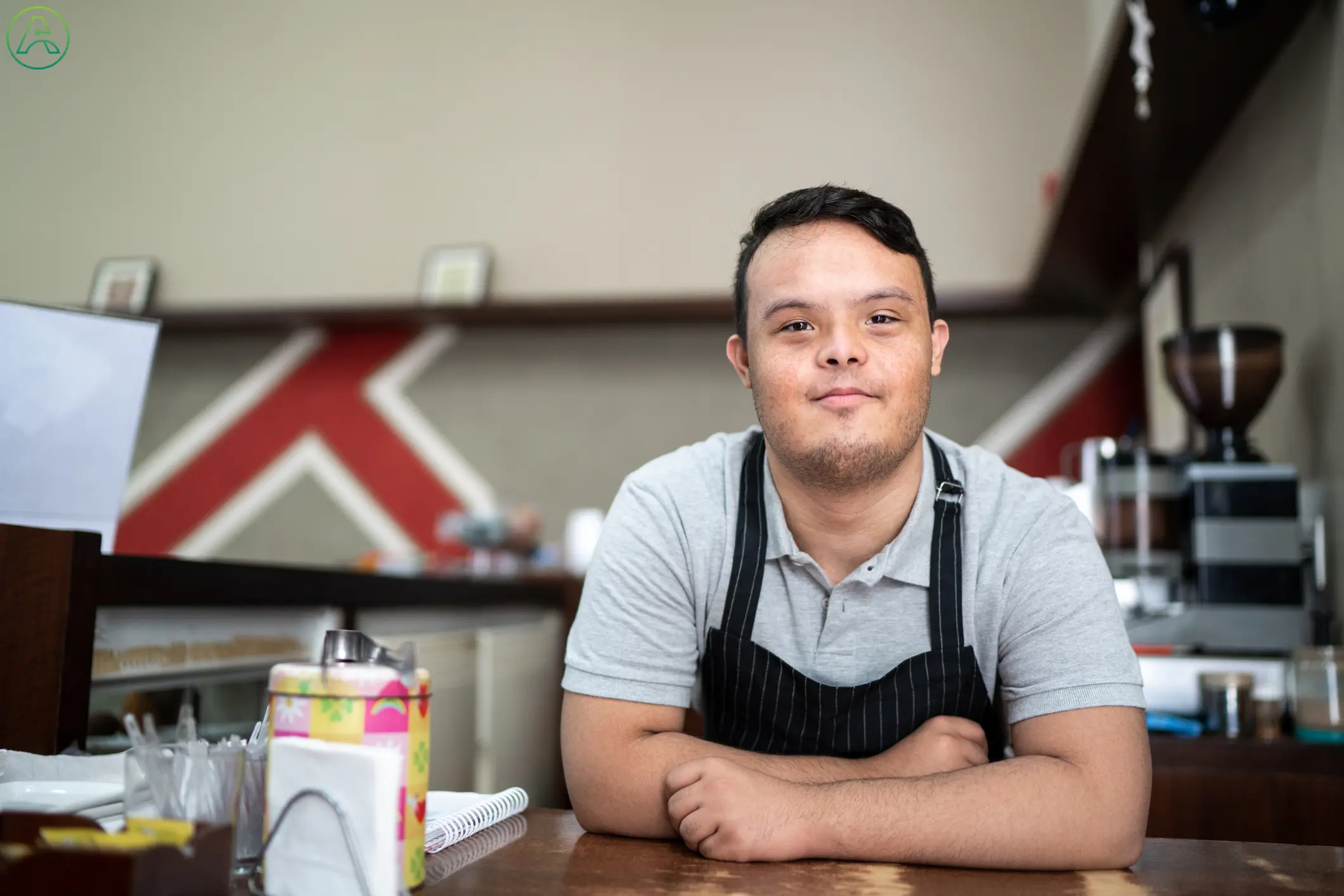 Wearing a gray tee shirt and a black striped apron, a smiling young mixed-race man with Down syndrome leans against the counter at his job as a coffee shop barista.