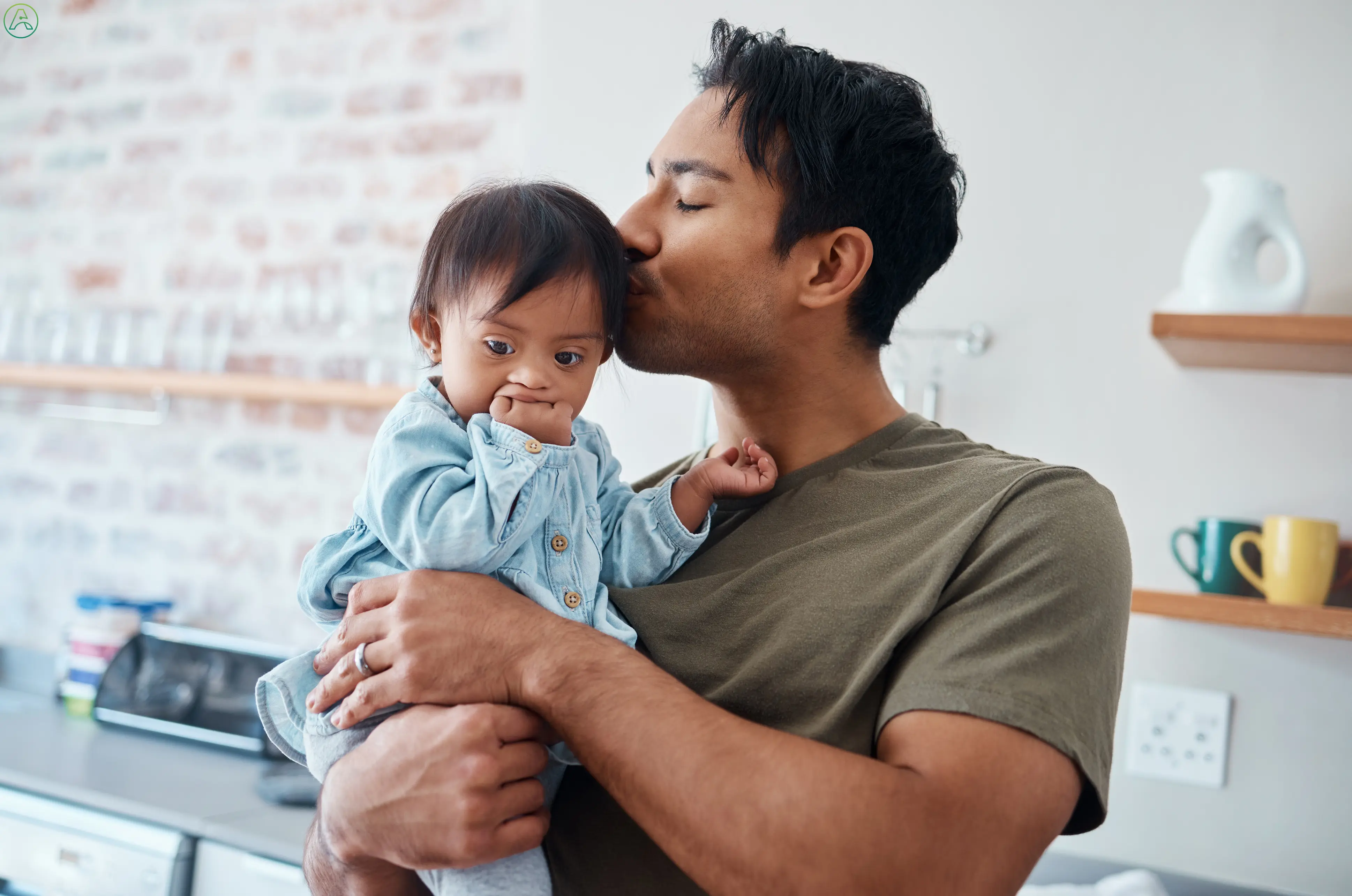 A young Latino man in a green tee shirt kisses his infant's head in their bright, modern kitchen. The baby, who has Down syndrome and is wearing a blue denim onesie, reaches for him with one hand while chewing on the other.