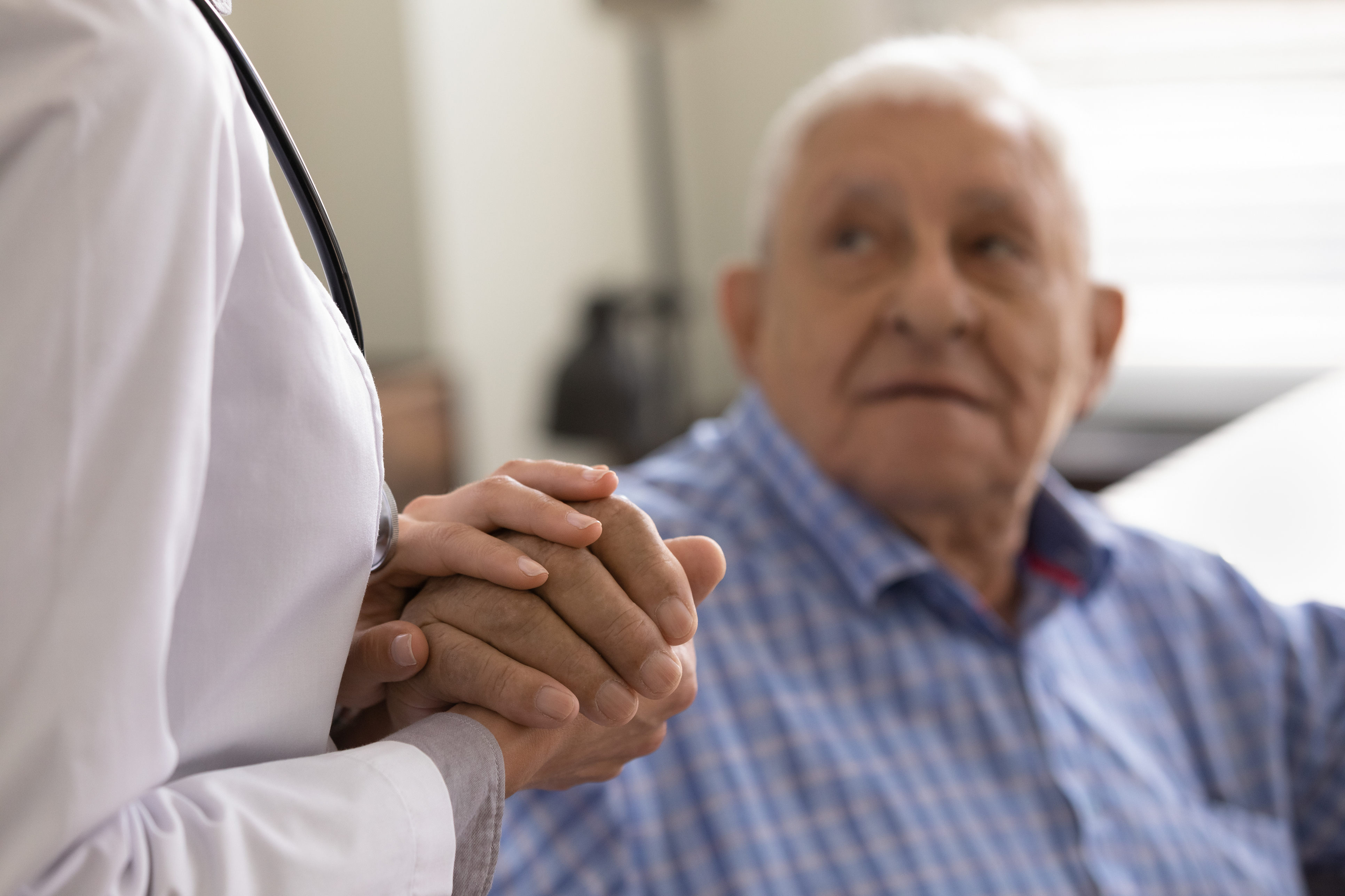 A white senior man sits and looks up at a doctor who holds his hand