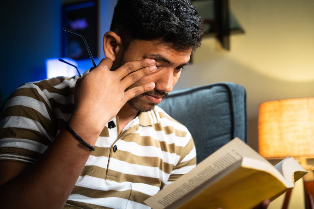 young Indian man rubs his eyes as he sits on a couch and tries to concentrate on reading a book
