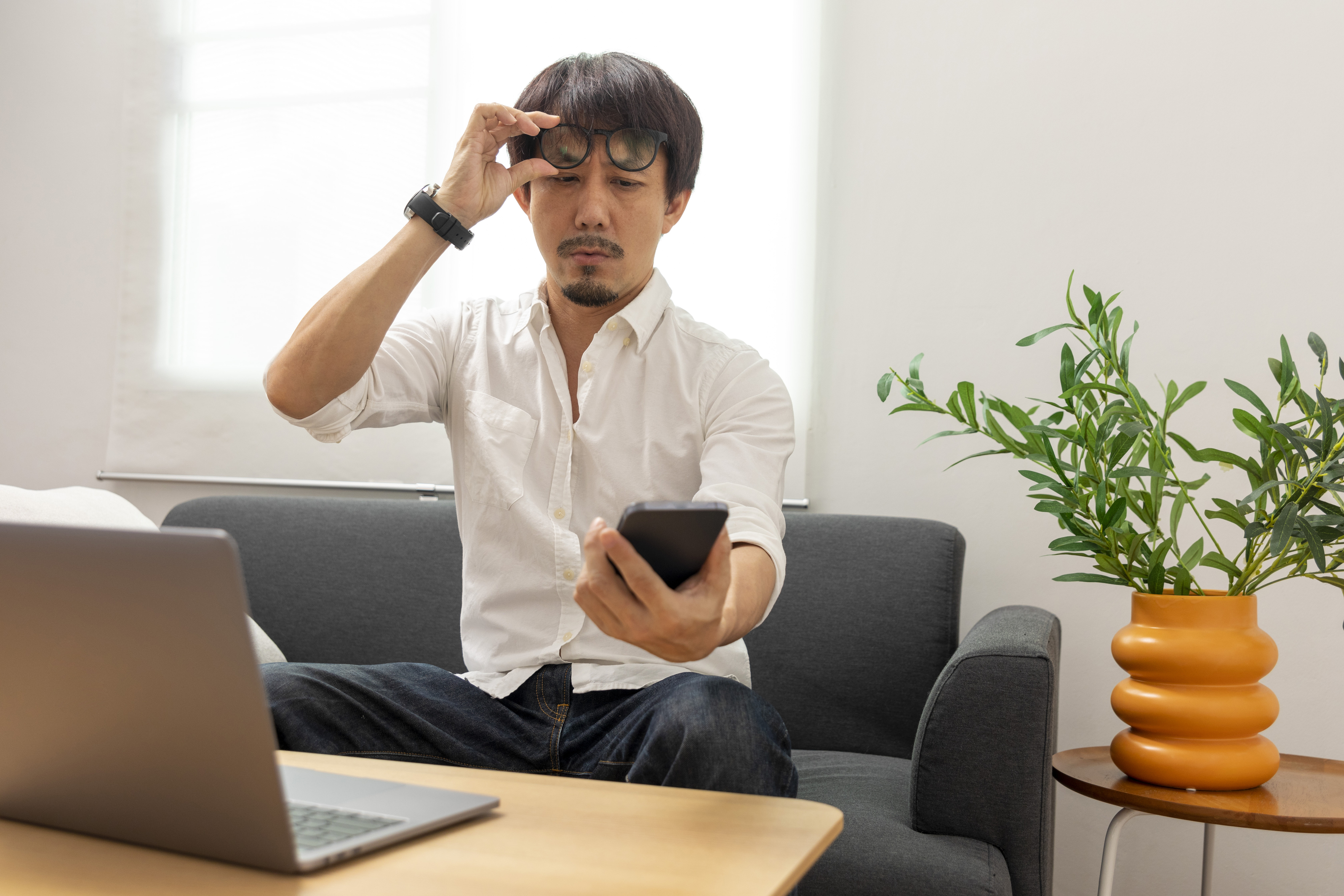 Asian man sits on a couch as he works from home, moving his glasses to adjust his eyes
